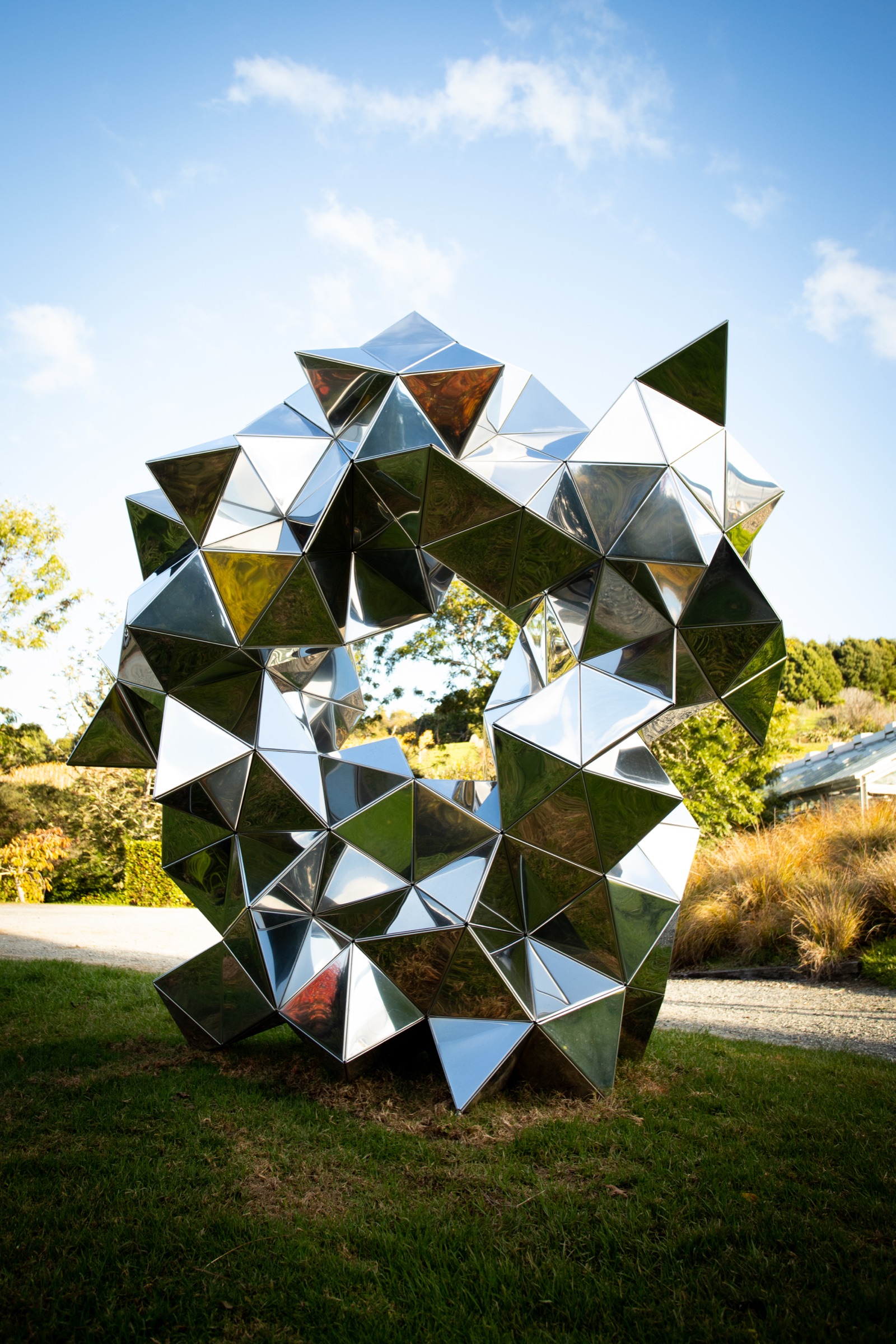 A large mirror-polished steel sculpture of faceted triangles on the lawn at Brick Bay Sculpture Trail, Matakana, reflecting the sky and trees, with tussock and the glass house roof behind.