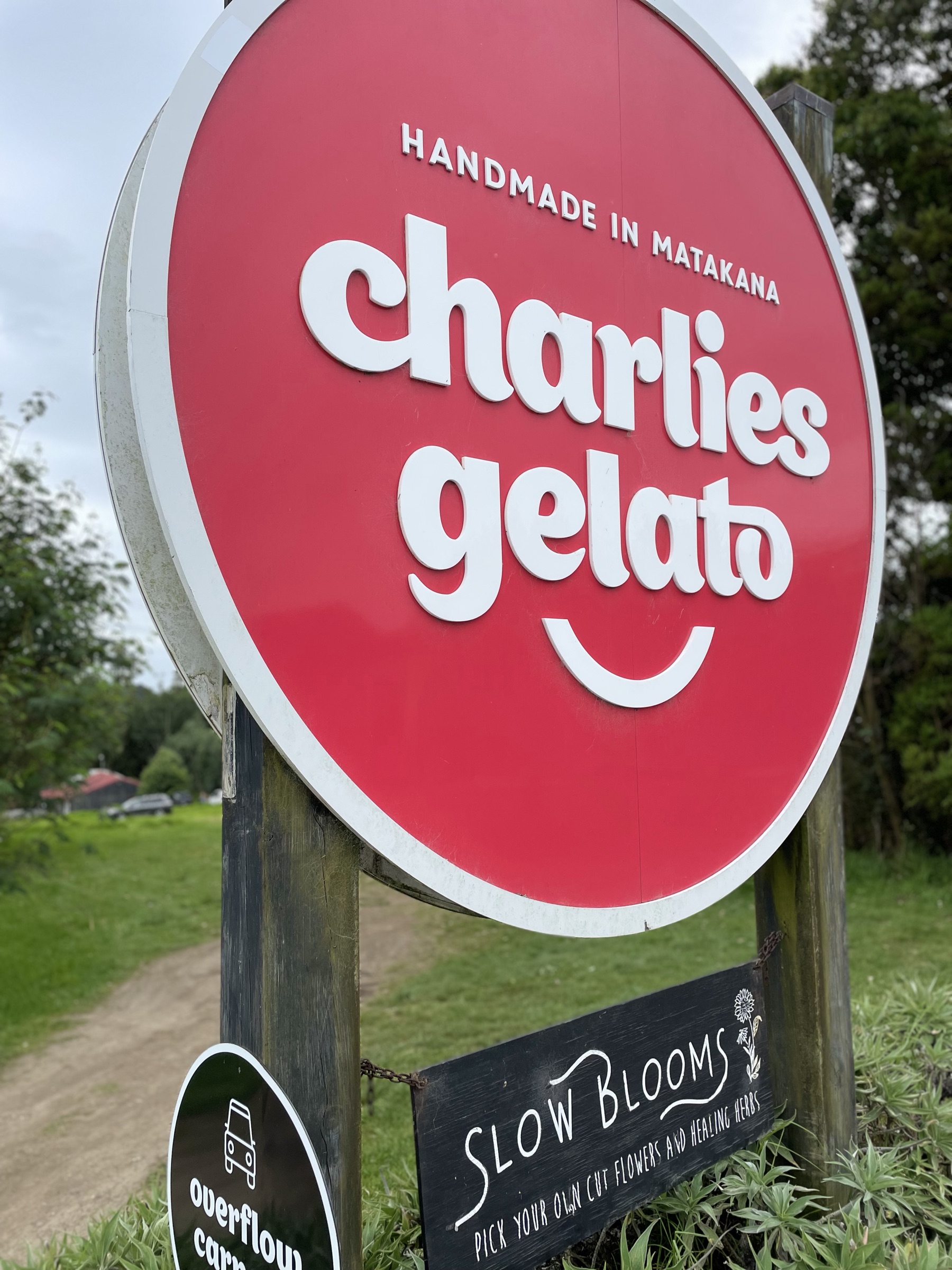 The round red Charlie's Gelato sign, handmade in Matakana, on timber posts at the gate, with the Slow Blooms pick-your-own flowers sign below and the paddock and trees behind.