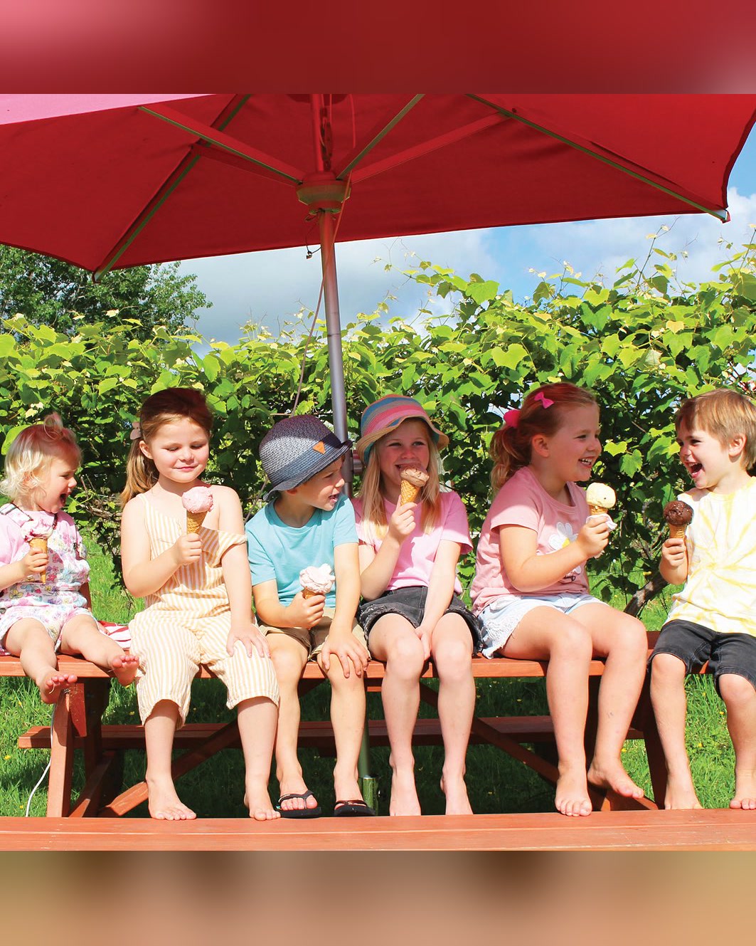 Six children in a row on a picnic bench under a red umbrella at Charlie's Gelato, Matakana, each with an ice cream cone, grapevines behind them.