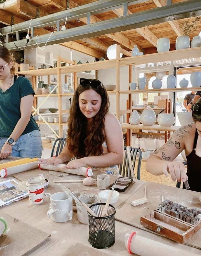Clay Friends studio, Matakana: a guest smiling as she rolls a slab of clay at a long bench, with tools, mugs, and clay stamps in front of her and shelves of finished pots behind.