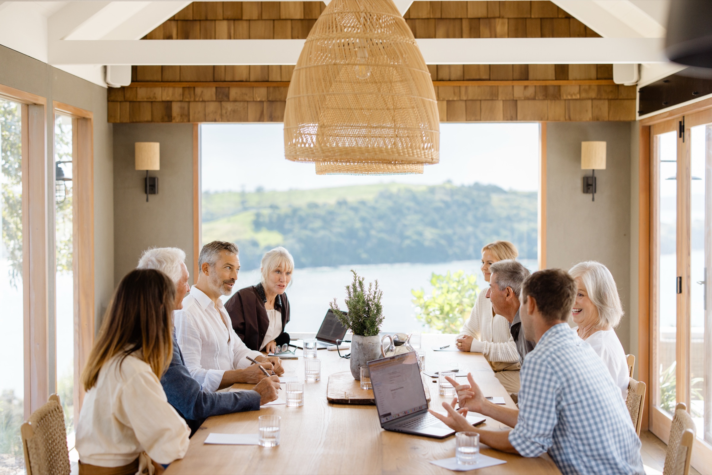 A board meeting at the Cookhouse, Bluestone Ridge: eight people around the oak table with laptops, water glasses, and rosemary in a stone pot, the rattan pendant above and the bay through the open wall behind.