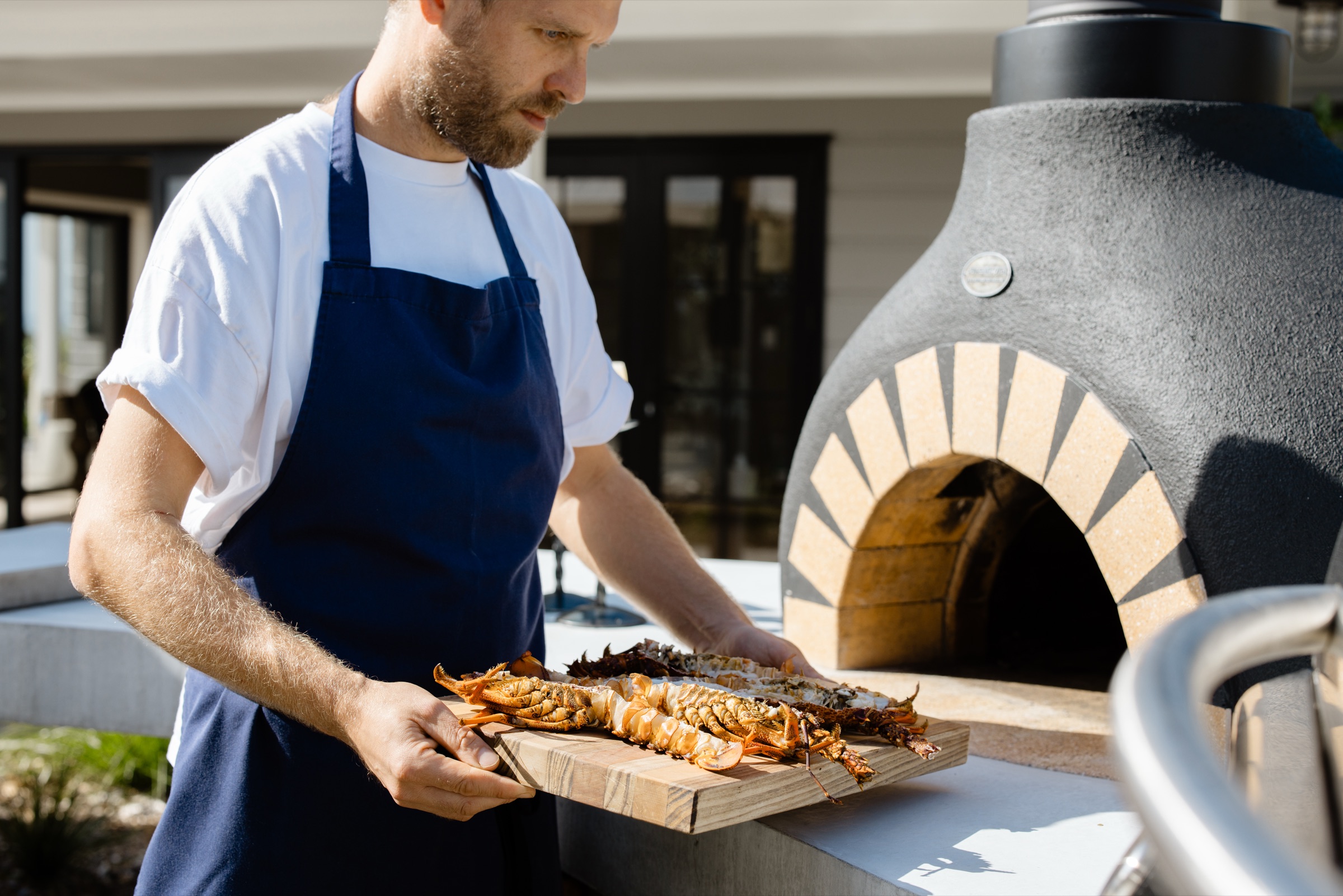 A chef in a navy apron carrying a board of crayfish to the wood-fired oven outside the Cookhouse at Bluestone Ridge.