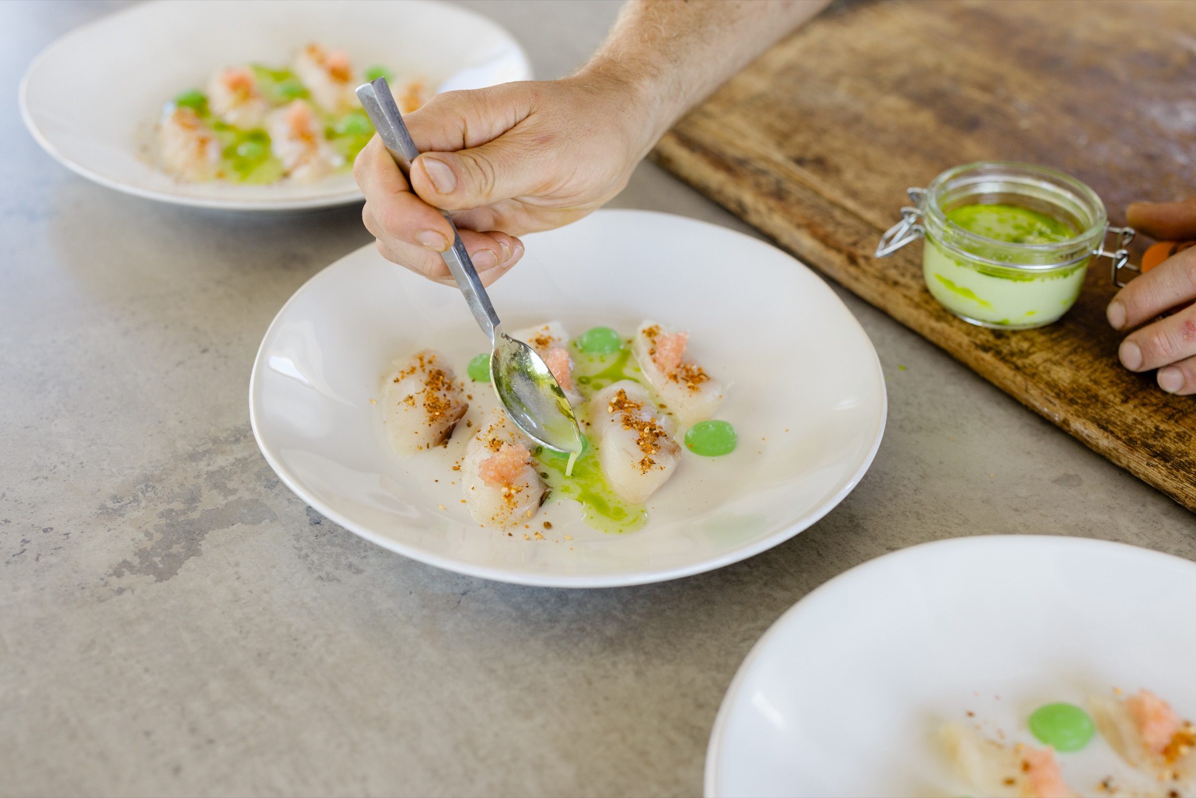 A chef spooning herb oil over cured fish with citrus on white plates at the Cookhouse bench, Bluestone Ridge.