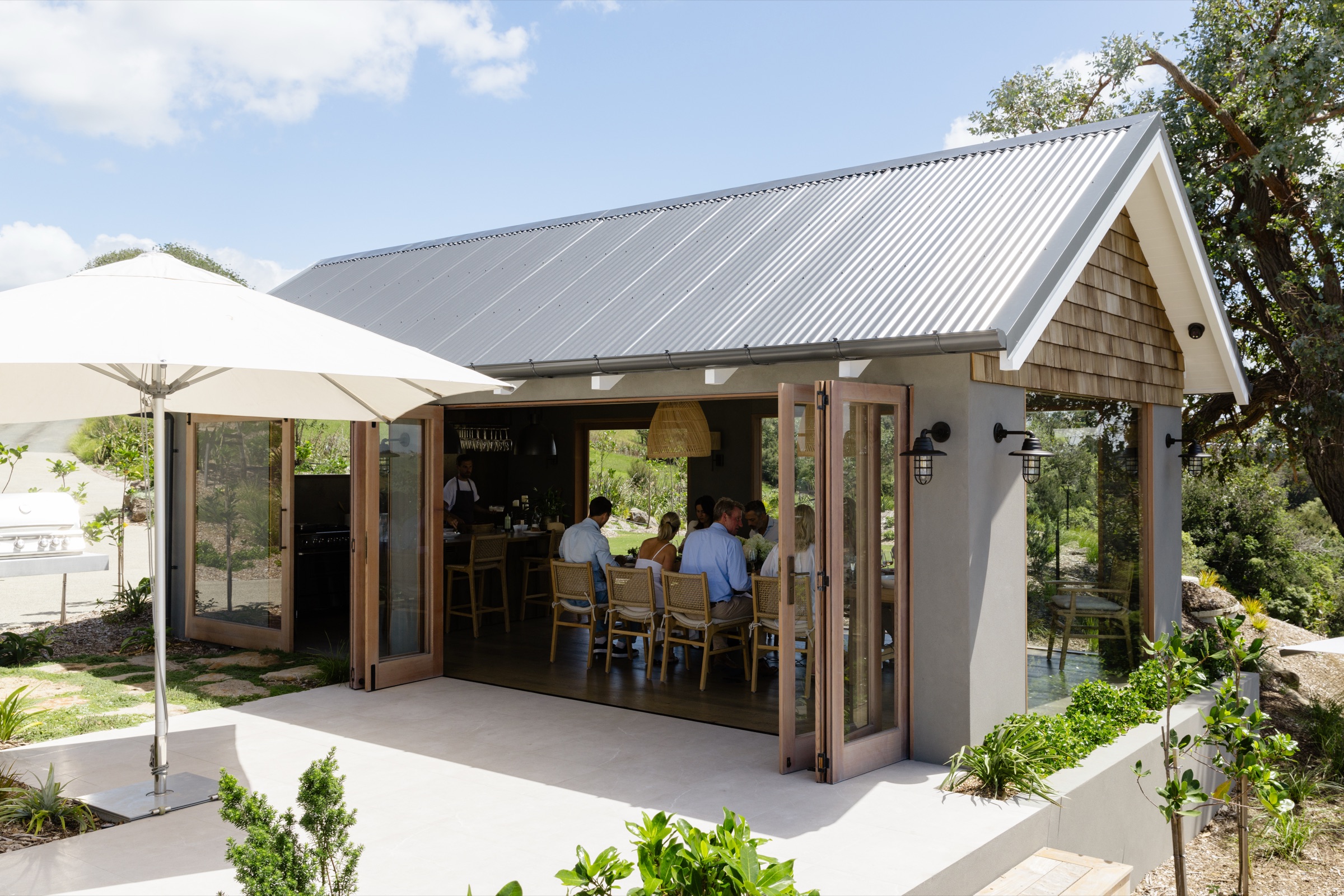 The Cookhouse at Bluestone Ridge from the terrace: corrugated roof and cedar shingle gable, the glass doors folded back, a chef at the bench and a party of guests at the oak table inside, an umbrella and the barbecue outside.