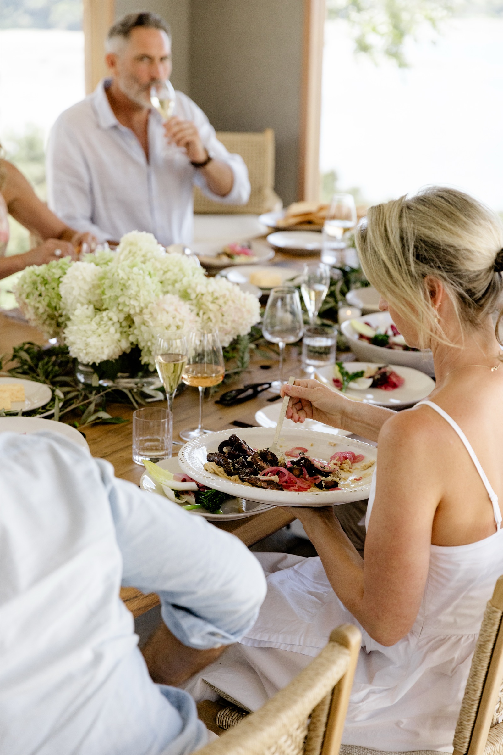 A guest in a white dress serving grilled octopus with pickled onion from a platter at the Cookhouse table, Bluestone Ridge, hydrangeas in the centre and a man with a glass of wine across the table.