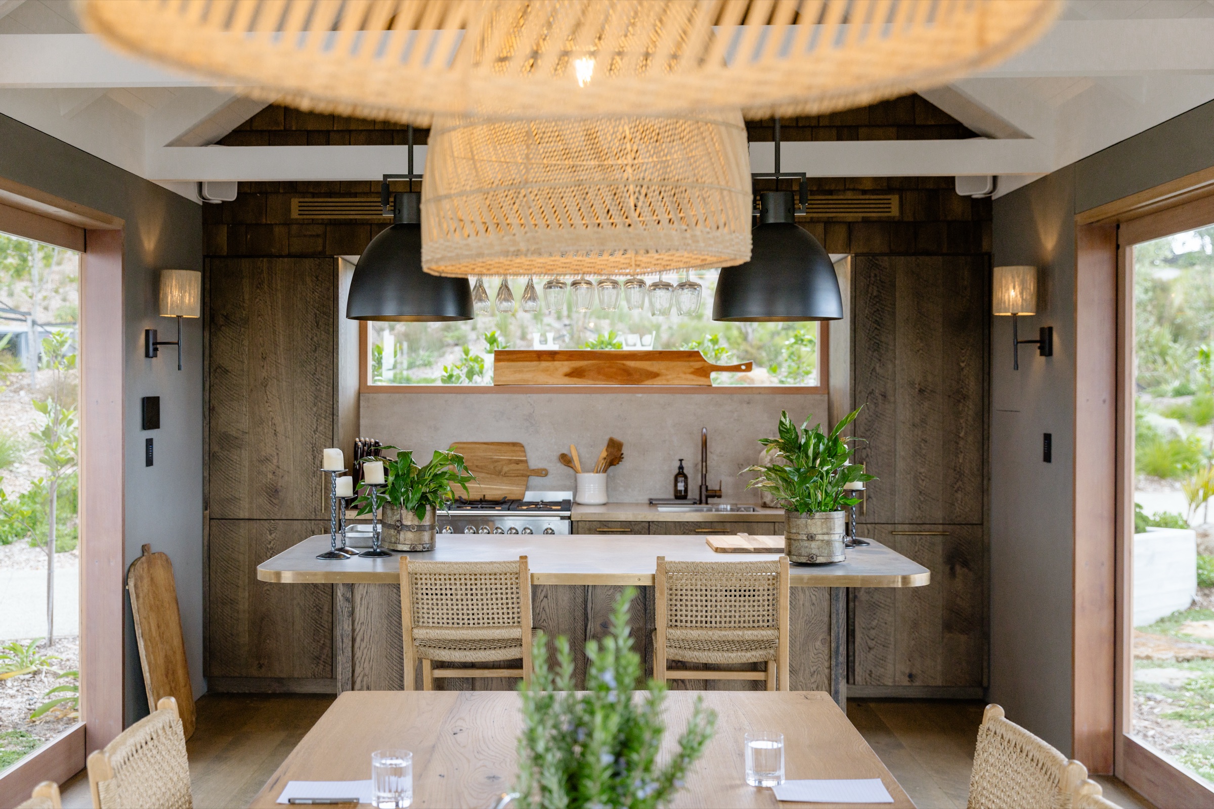 The Cookhouse kitchen at Bluestone Ridge: a concrete-topped island with brass edge and two rattan stools, the gas range and concrete splashback behind, glasses hanging under black and rattan pendants, and dark oak cabinetry.