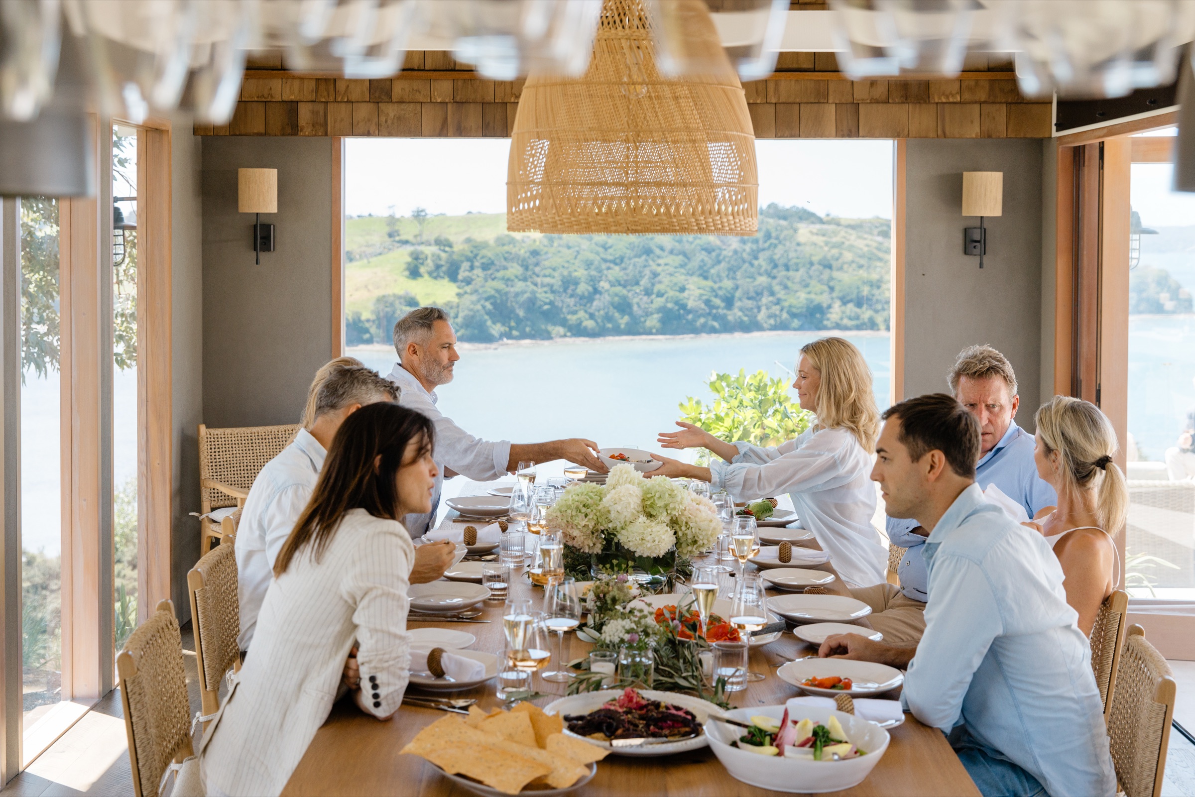 Lunch at the Cookhouse, Bluestone Ridge: guests passing dishes down the oak table under the rattan pendant, hydrangeas and platters between them, and the inlet and green hills through the open wall behind.