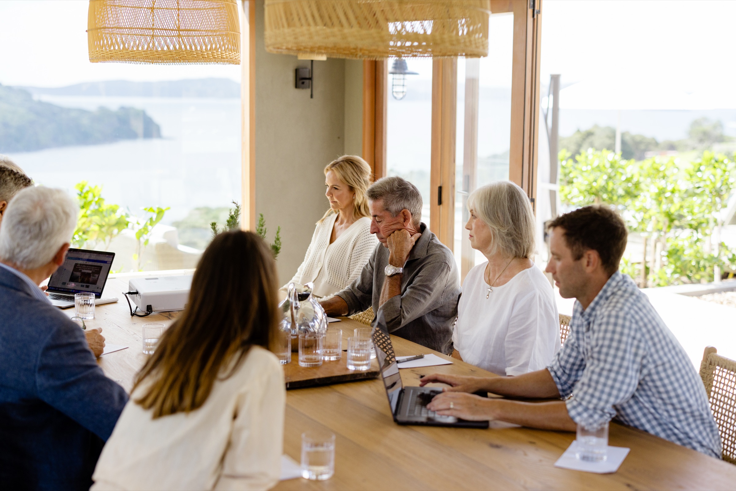 A meeting at the Cookhouse table, Bluestone Ridge: colleagues with laptops and a projector, the bifold doors open to the water and hills.