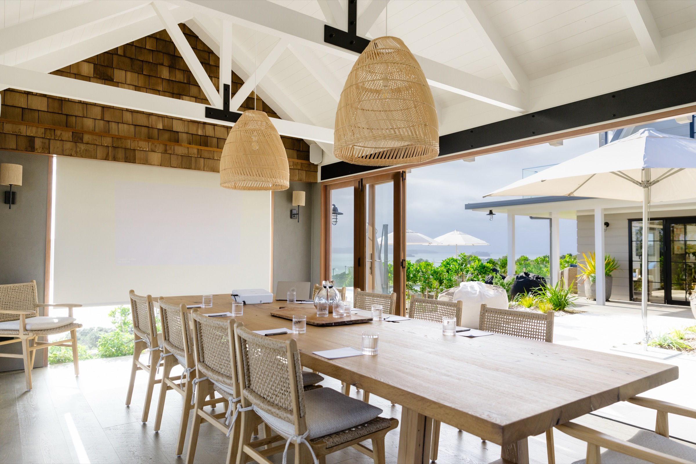 The Cookhouse at Bluestone Ridge set for a meeting: the oak table with water glasses and notepads, a projector and screen at the end, rattan pendants under the white trusses, and the doors open to the terrace and the Gulf.