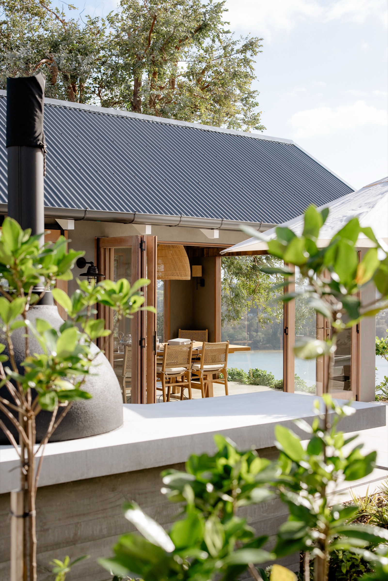 The wood-fired oven on its concrete bench outside the Cookhouse at Bluestone Ridge, the glass doors open to the oak table and chairs, and the water beyond.
