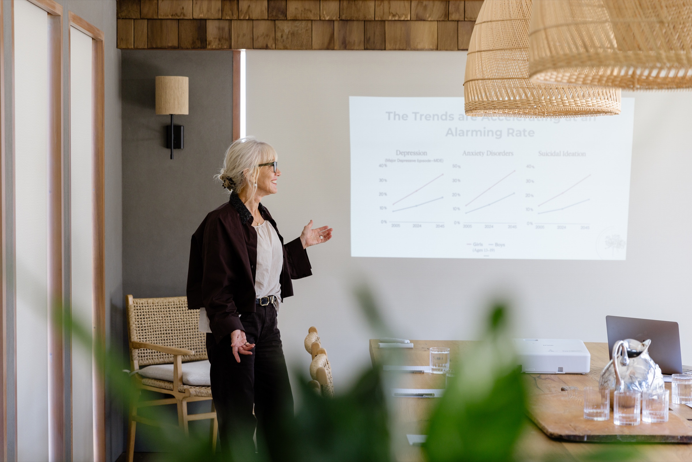 A presenter at the Cookhouse, Bluestone Ridge, standing beside charts projected onto the wall screen, rattan pendants above the table.