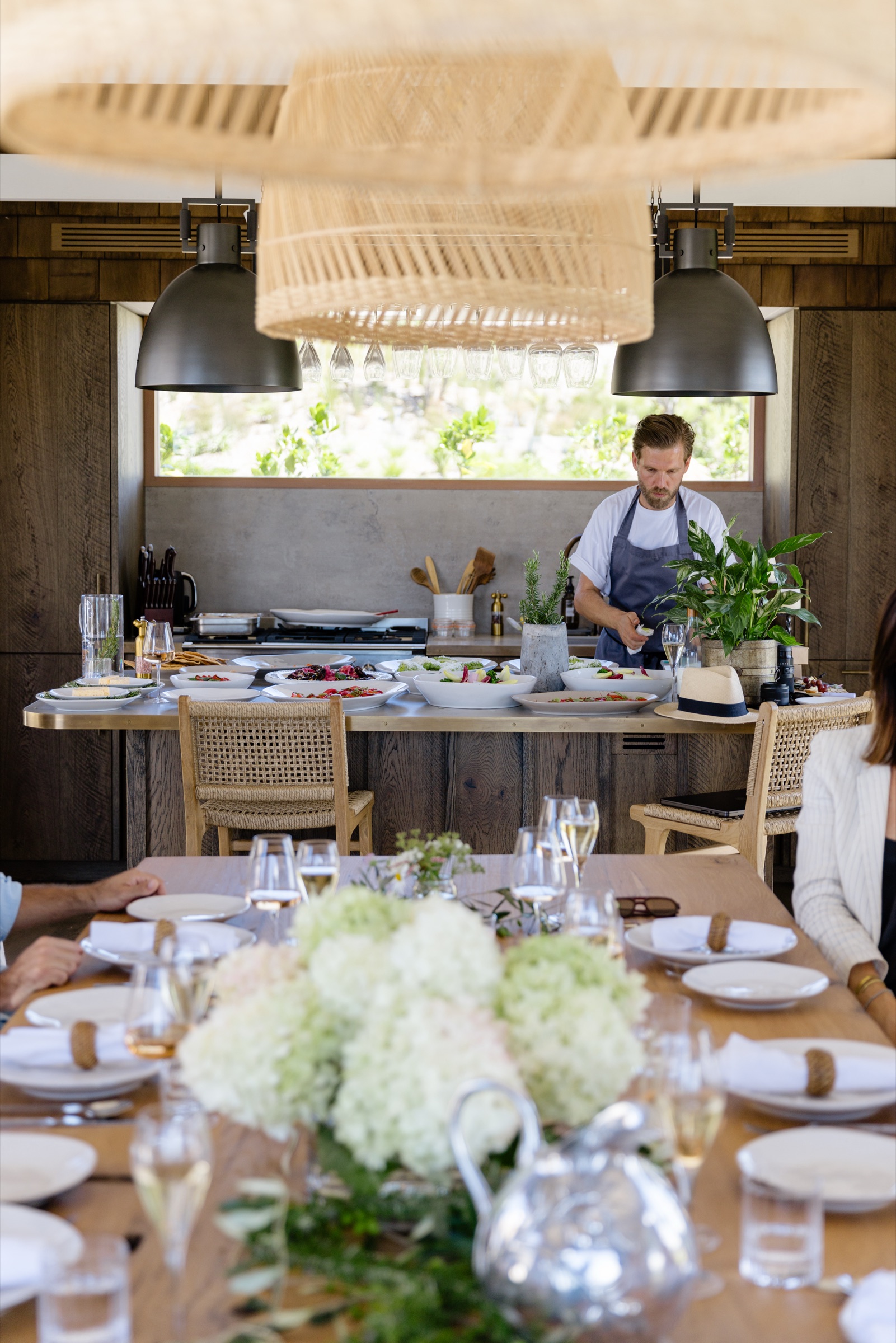 A private chef in a grey apron plating lunch at the Cookhouse island at Bluestone Ridge, platters of salads and seafood lined along the brass-edged bench, glasses hanging under the rattan pendants, and the set oak table with hydrangeas in the foreground.