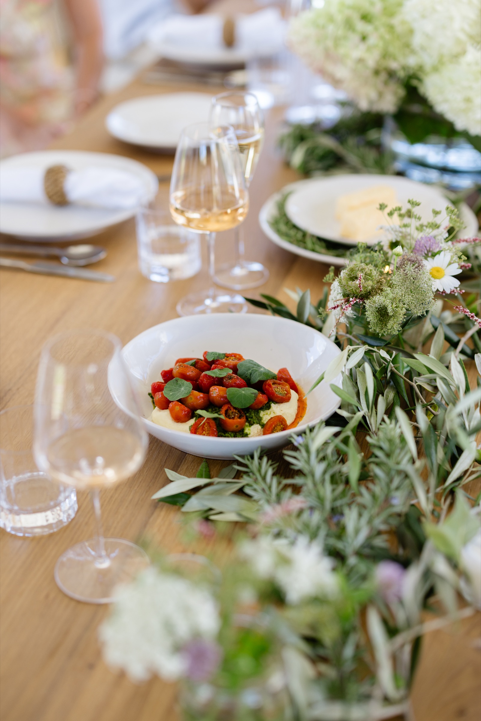 A bowl of roasted cherry tomatoes with nasturtium leaves on whipped feta on the Cookhouse table, Bluestone Ridge, among rosemary, olive branches, hydrangeas, and glasses of rosé.