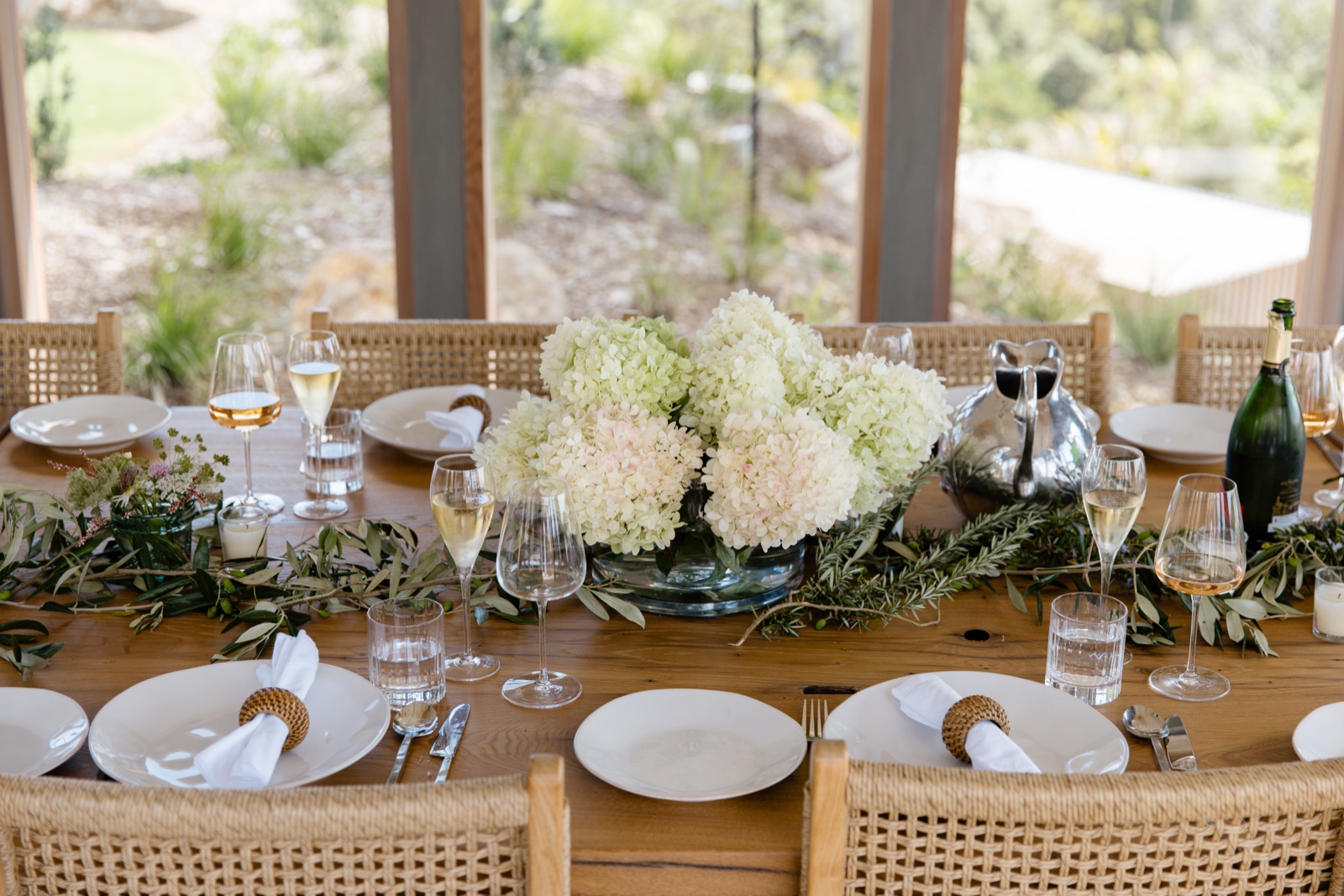 The Cookhouse table at Bluestone Ridge set for a celebration: white and blush hydrangeas in a glass bowl, olive and rosemary branches, rattan napkin rings, champagne, and the garden through the glass.