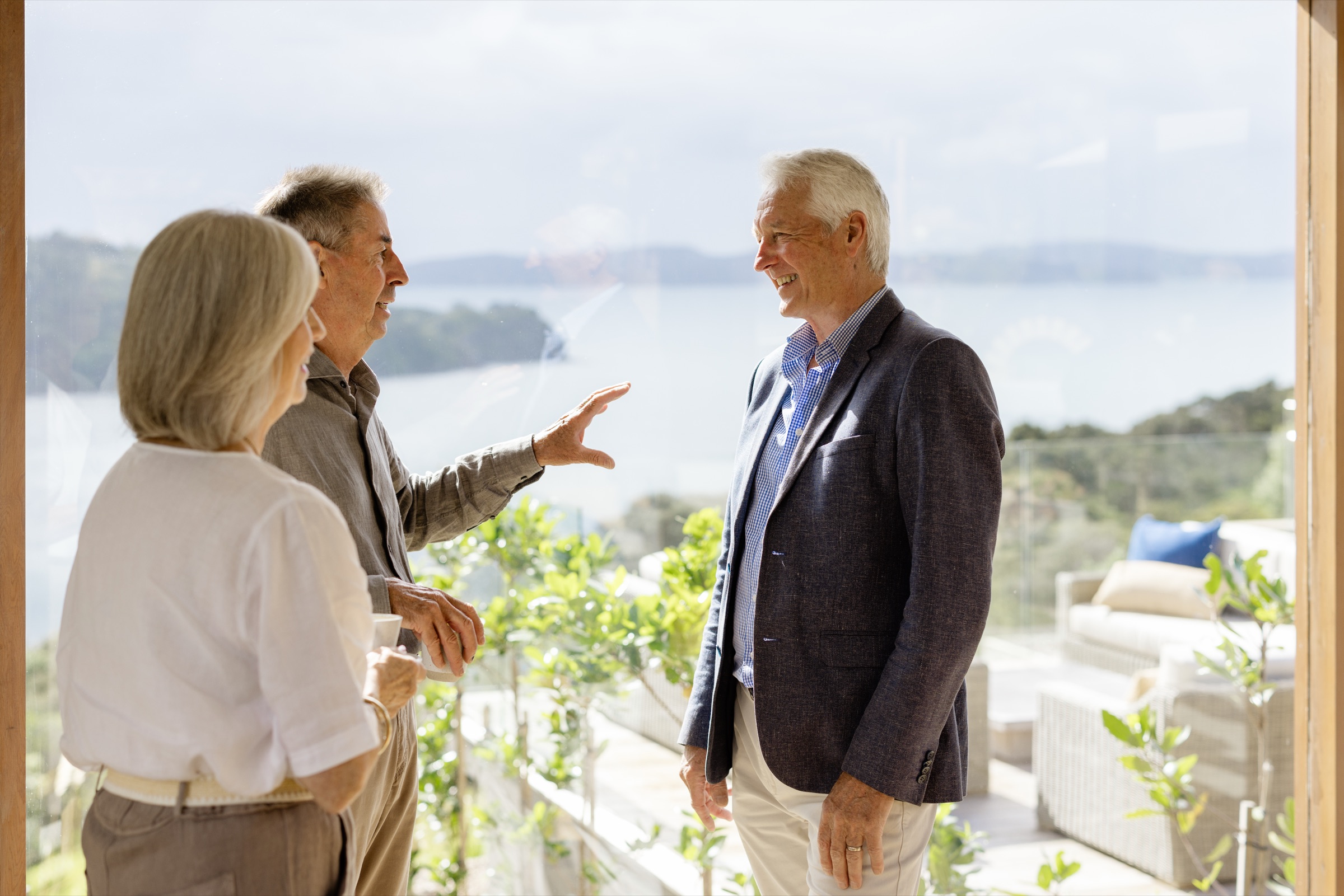 Three colleagues talking at the open doors of the Cookhouse, Bluestone Ridge, one gesturing to the bay and the headland beyond, the terrace sofas below.