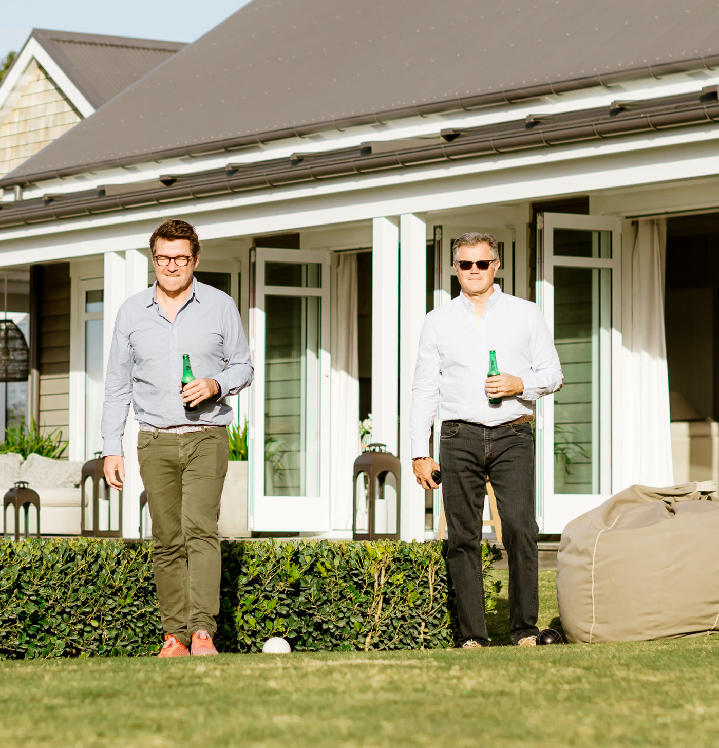 Two executives with beers playing lawn games on the grass in front of the Main House verandah at Bluestone Main Estate, beanbags and an umbrella nearby.