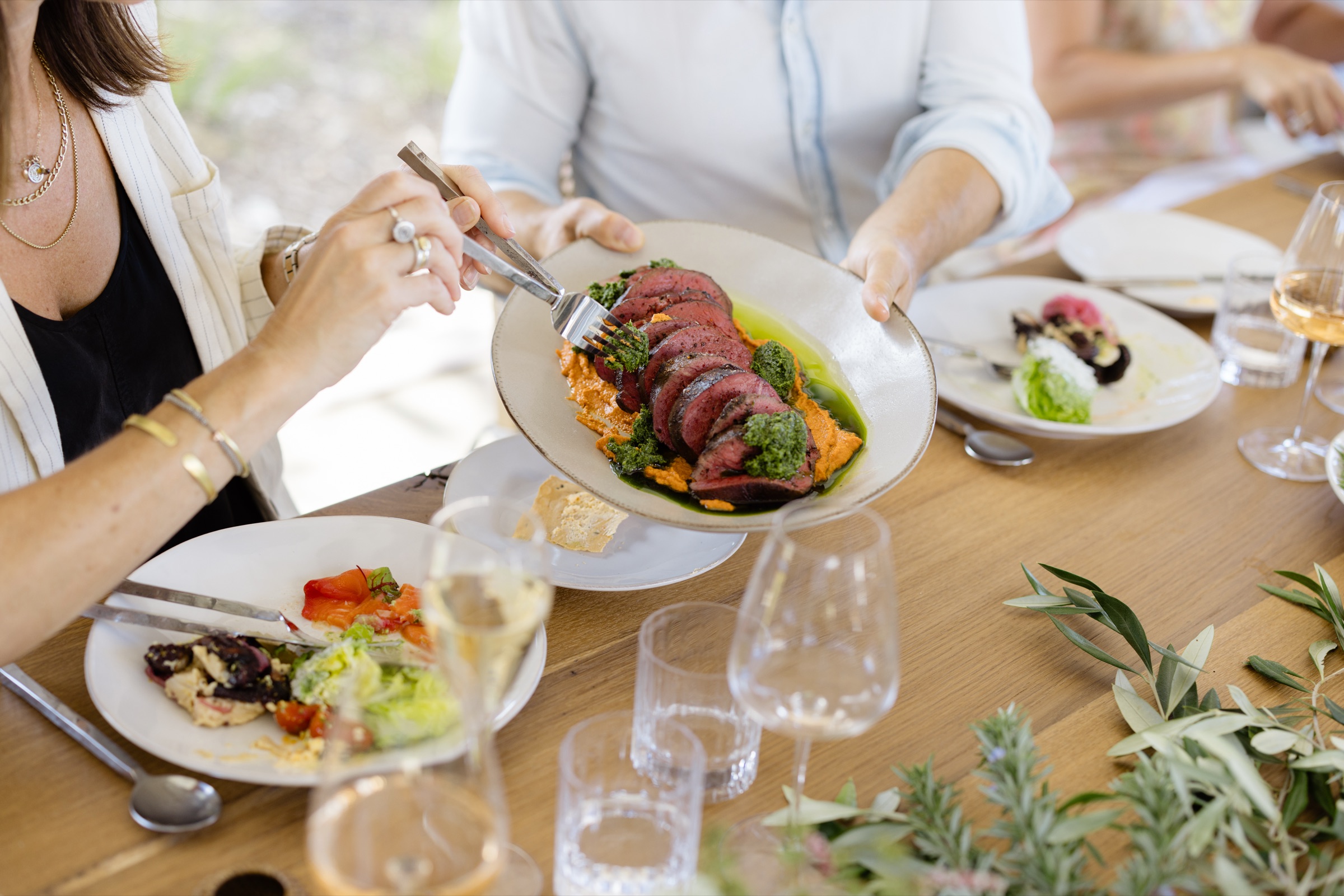 Lunch at Bluestone Estate: a platter of sliced beef with romesco and salsa verde passed along the oak table, a guest serving herself, wine glasses and olive branches on the table, no faces in frame.