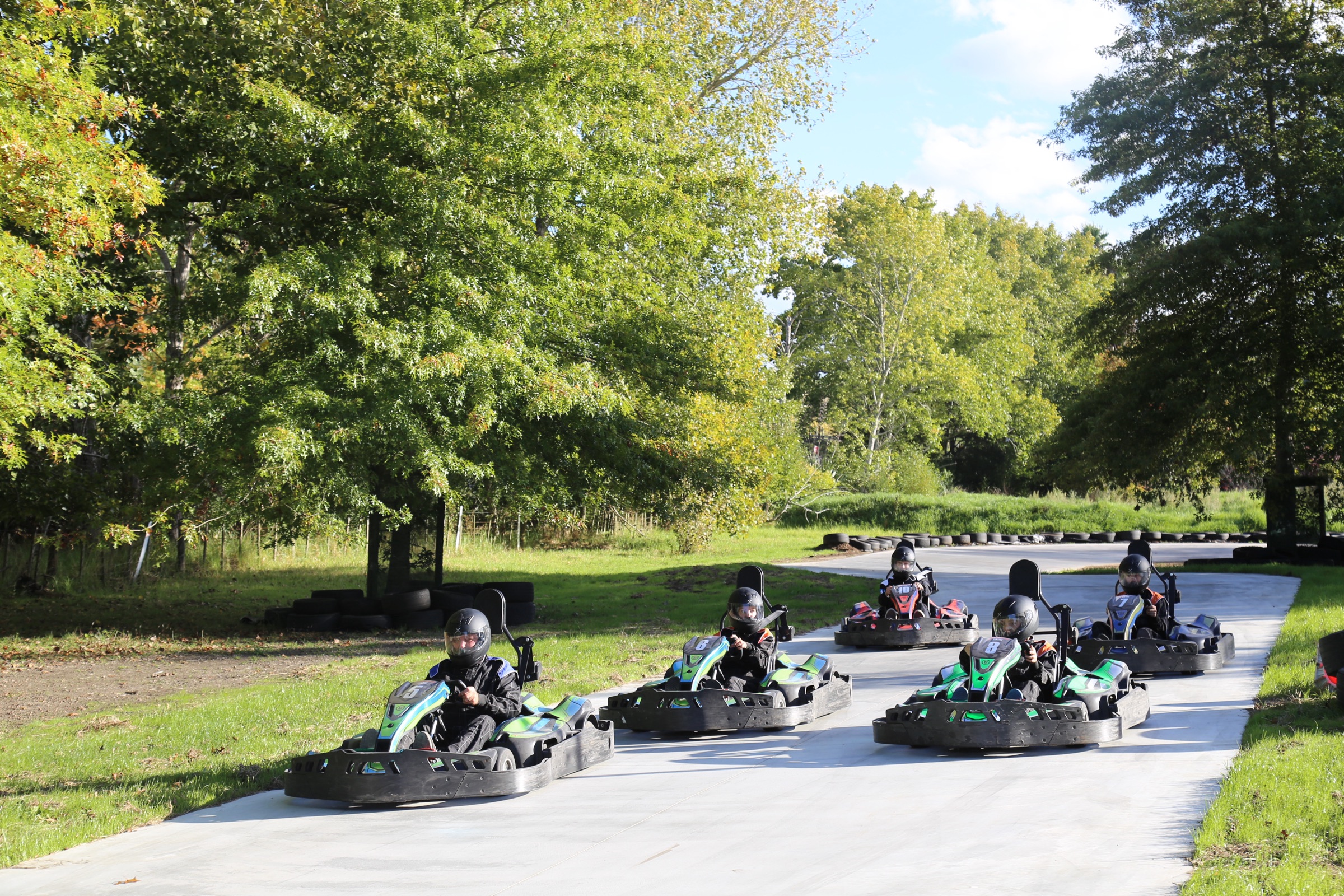 Five electric karts in a pack on the concrete track at Matakana Country Park, drivers in helmets and race suits, tyre walls and big trees around the circuit.