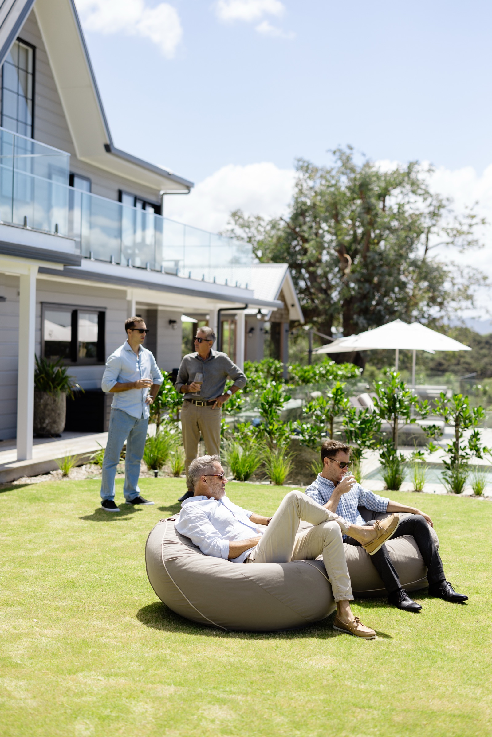 Four men with drinks on the lawn at the Farmhouse, Bluestone Ridge: two on a beanbag, two standing by the verandah, the pool umbrellas behind.