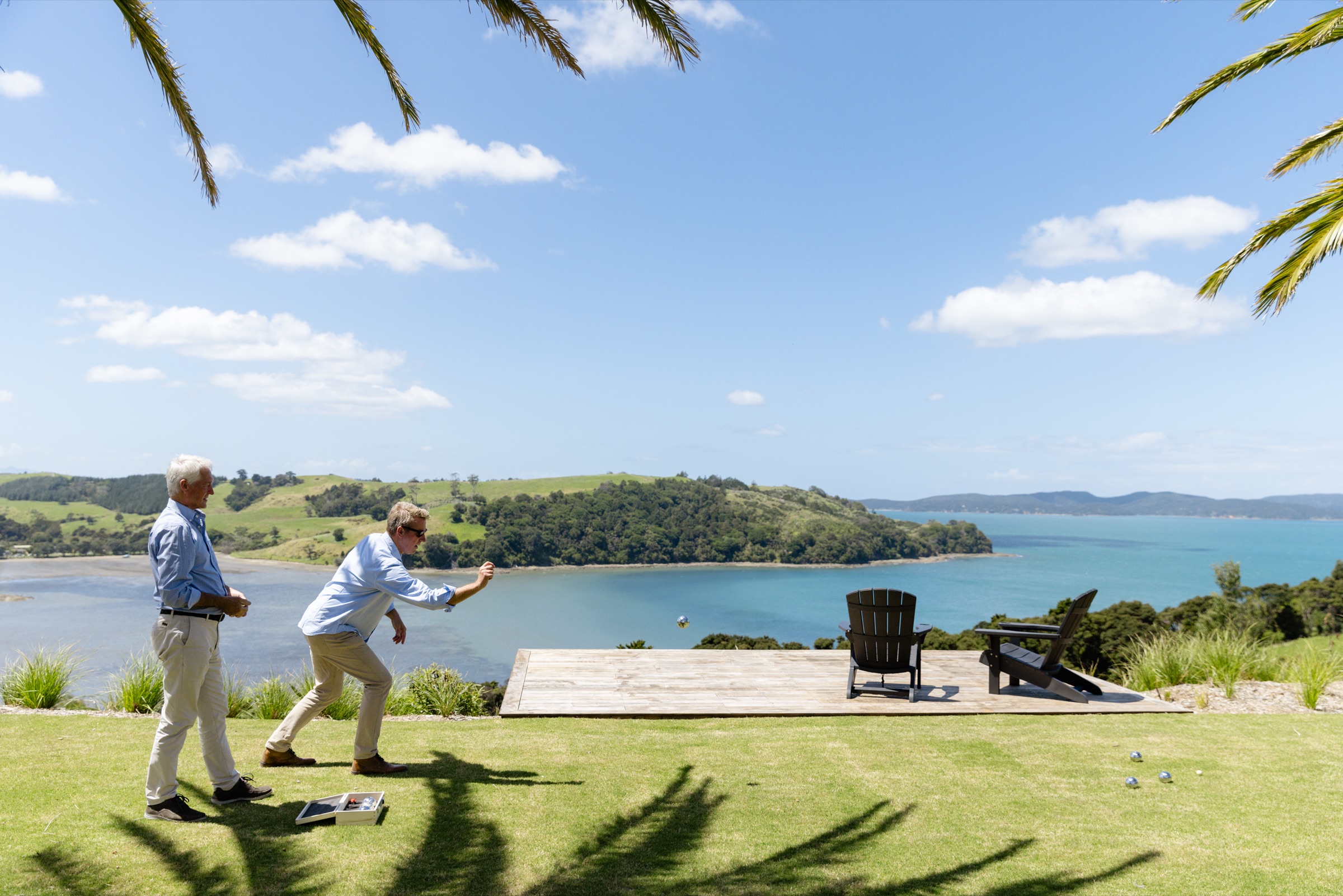 Two men playing pétanque on the lawn at the Farmhouse, Bluestone Ridge, palm fronds above, two Adirondack chairs on a platform, and the bay and green hills beyond.