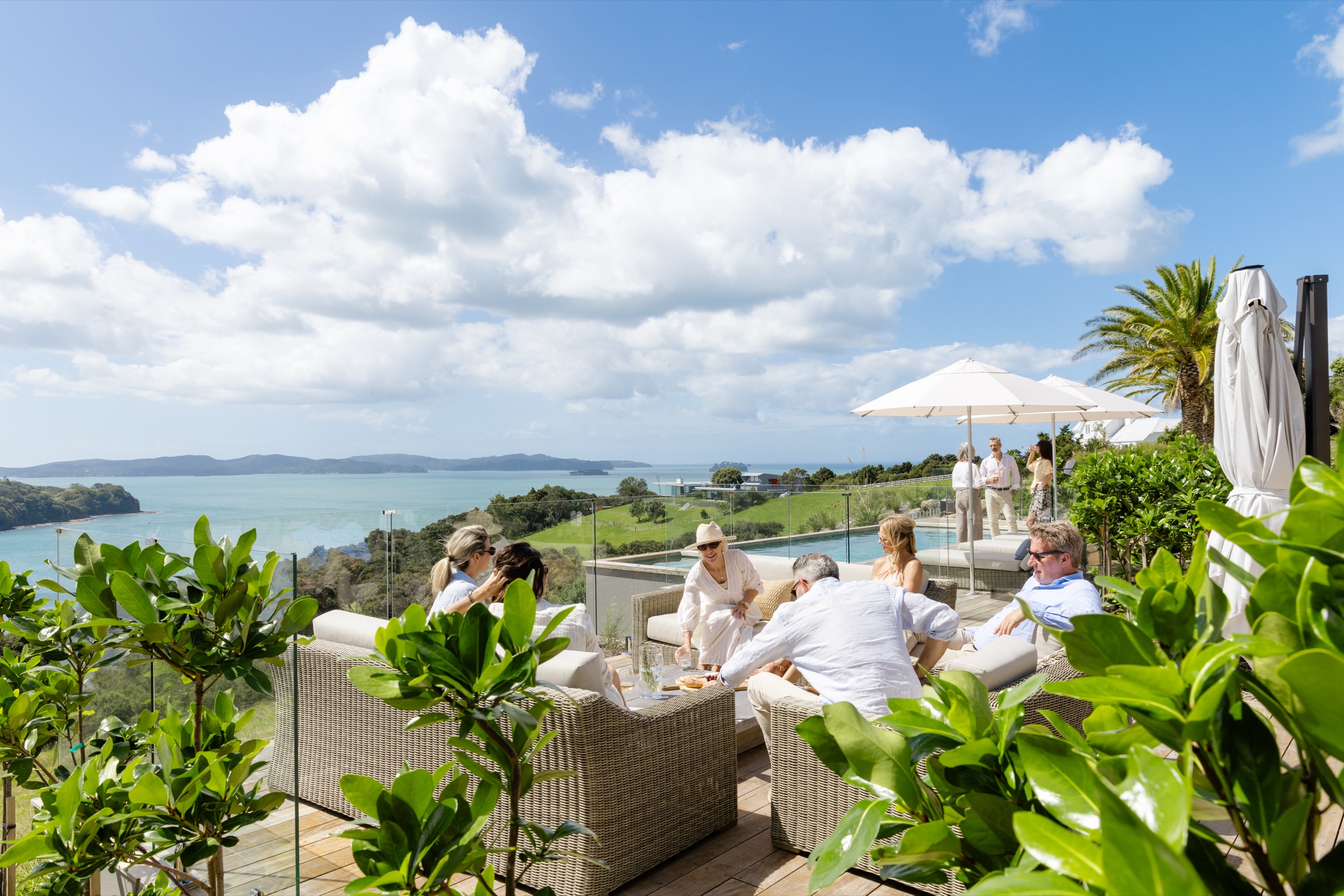 Guests on the rattan sofas beside the Farmhouse pool at Bluestone Ridge, umbrellas up, the infinity pool behind them and the Hauraki Gulf and its islands beyond the paddocks.