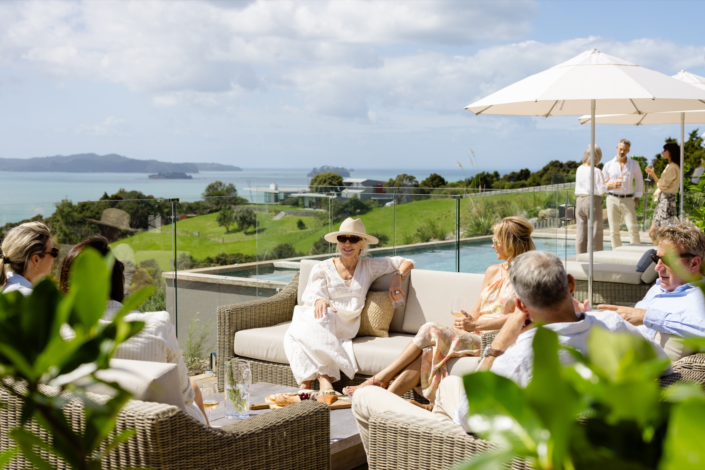 A group on the pool terrace at the Farmhouse, Bluestone Ridge: a woman in a white dress and sun hat on the sofa, a platter on the table, others standing by the pool under white umbrellas, the Gulf behind.