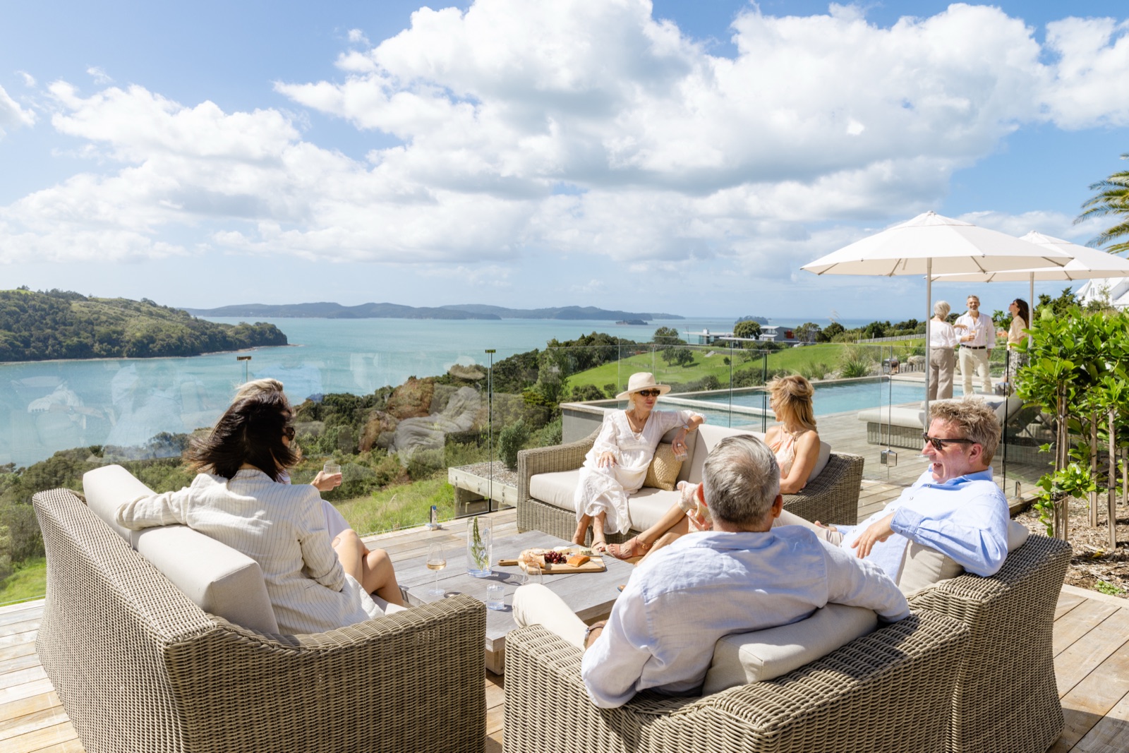 A team on the rattan sofas of the pool terrace at the Farmhouse, Bluestone Ridge, wine and a grazing board on the table, the infinity pool and umbrellas behind, and Kawau Bay and its islands beyond.