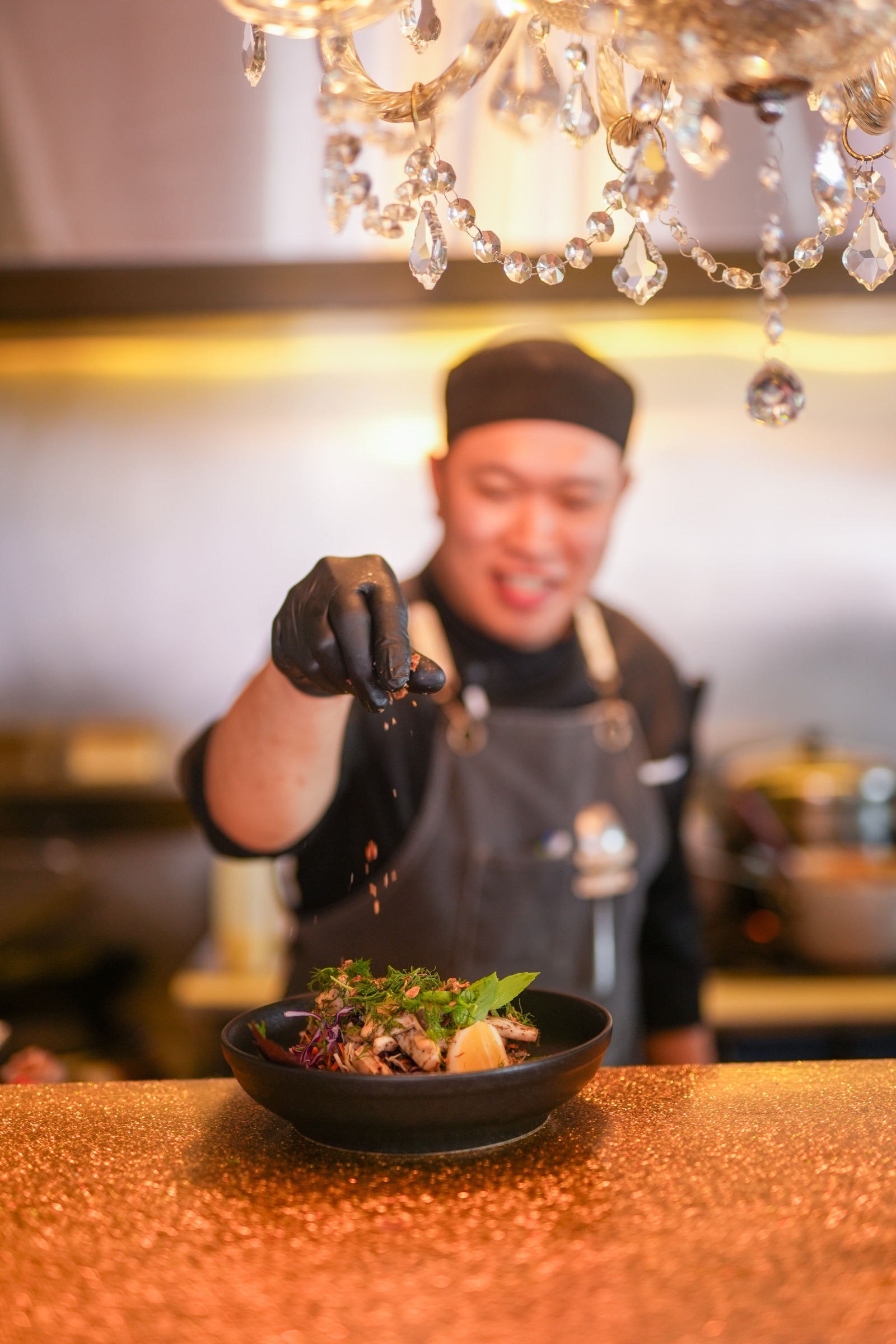 A chef at Jin Jin, Matakana, sprinkling the finishing touch over a bowl of squid salad on the gold counter, a crystal chandelier above and the kitchen soft behind.