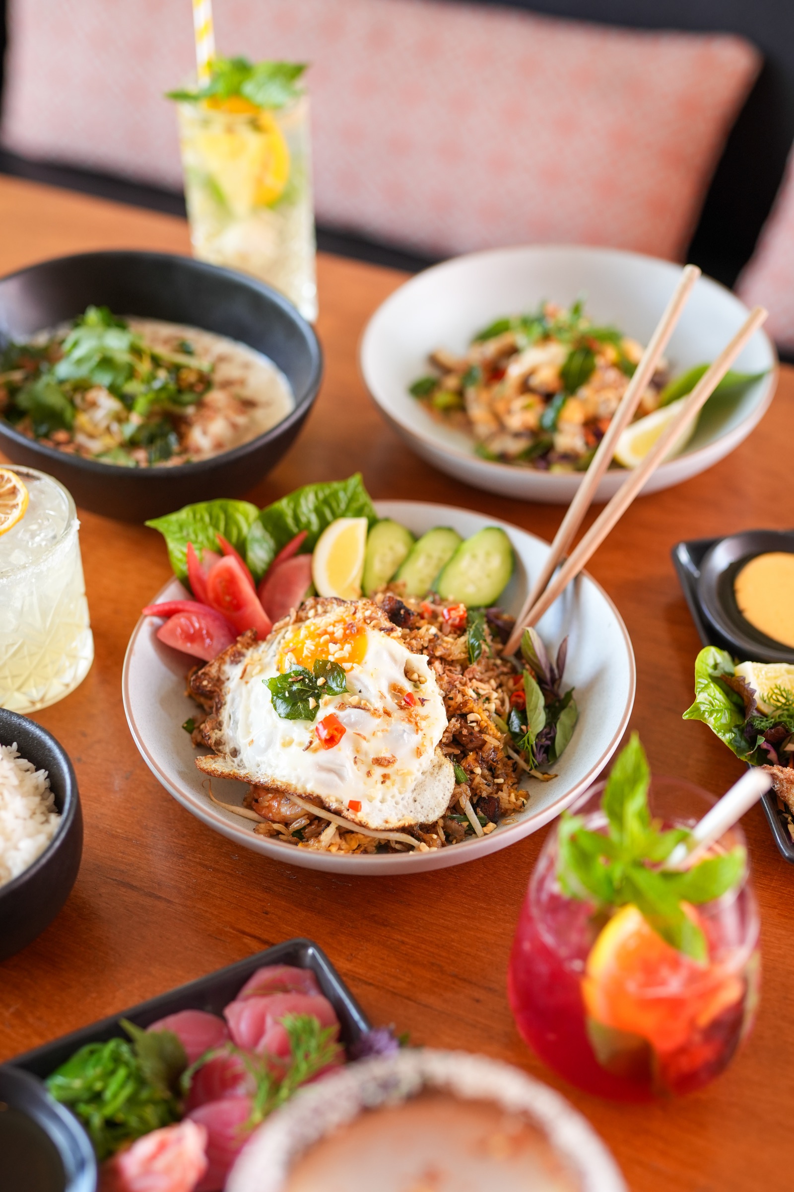 A table at Jin Jin, Matakana: fried rice topped with a fried egg, chilli, and herbs, a bowl of soup, a salad with chopsticks, a tray of sashimi, a bowl of rice, and two cocktails on the timber table.