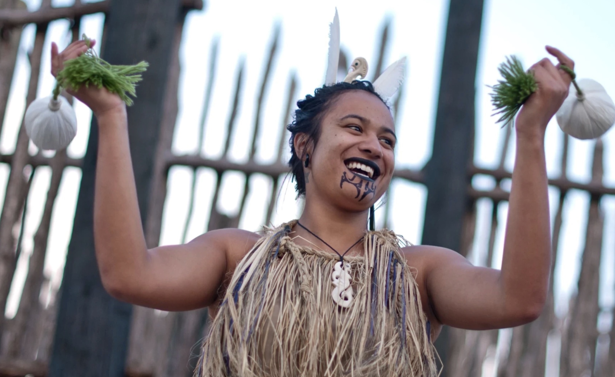 A Māori performer with moko kauae smiling mid-song, poi raised in both hands, in a flax bodice and feather headpiece before a timber palisade.