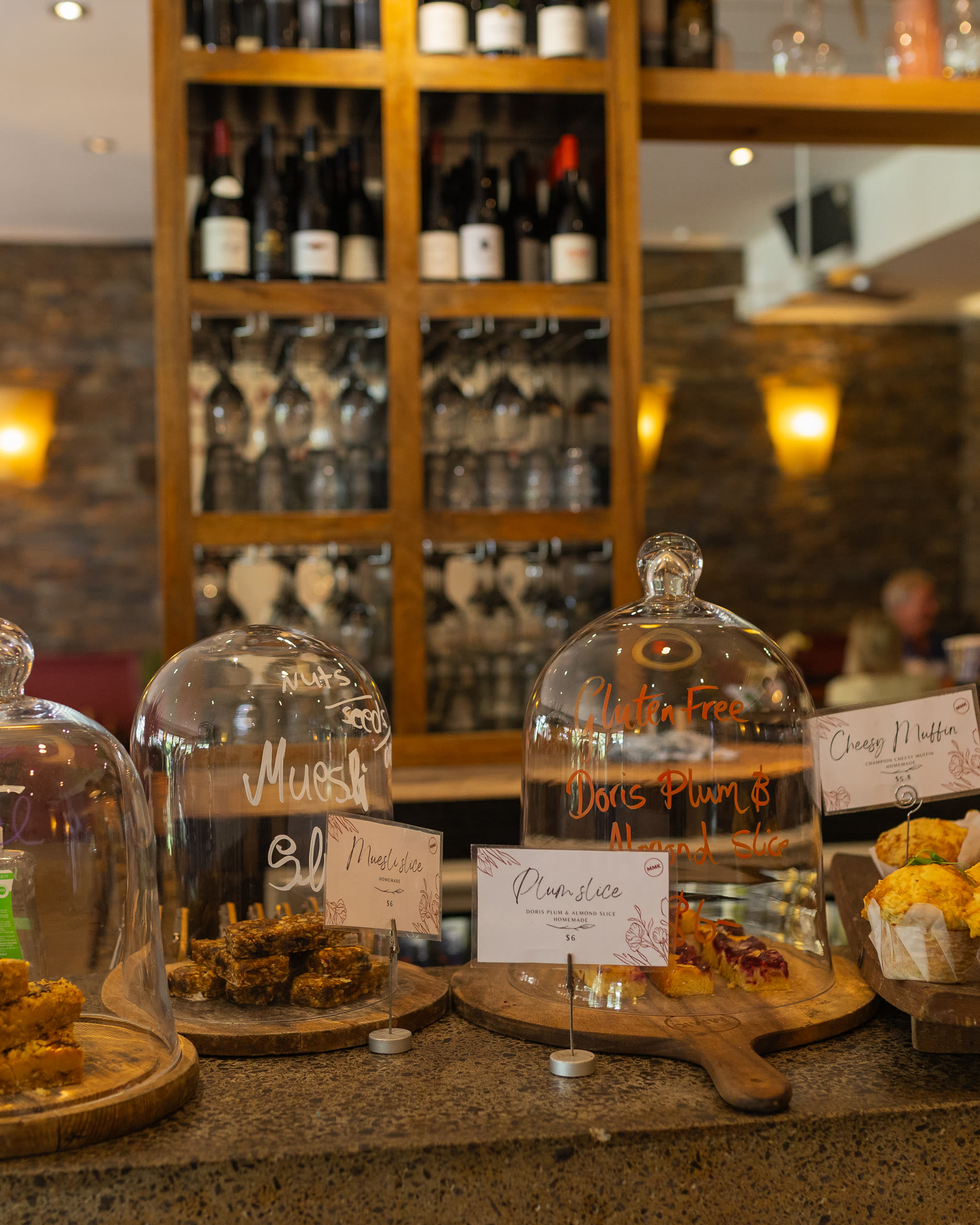 The counter at Matakana Market Kitchen: homemade slices and cheesy muffins under glass cloches with handwritten labels, the wine shelves and wall lamps soft behind.