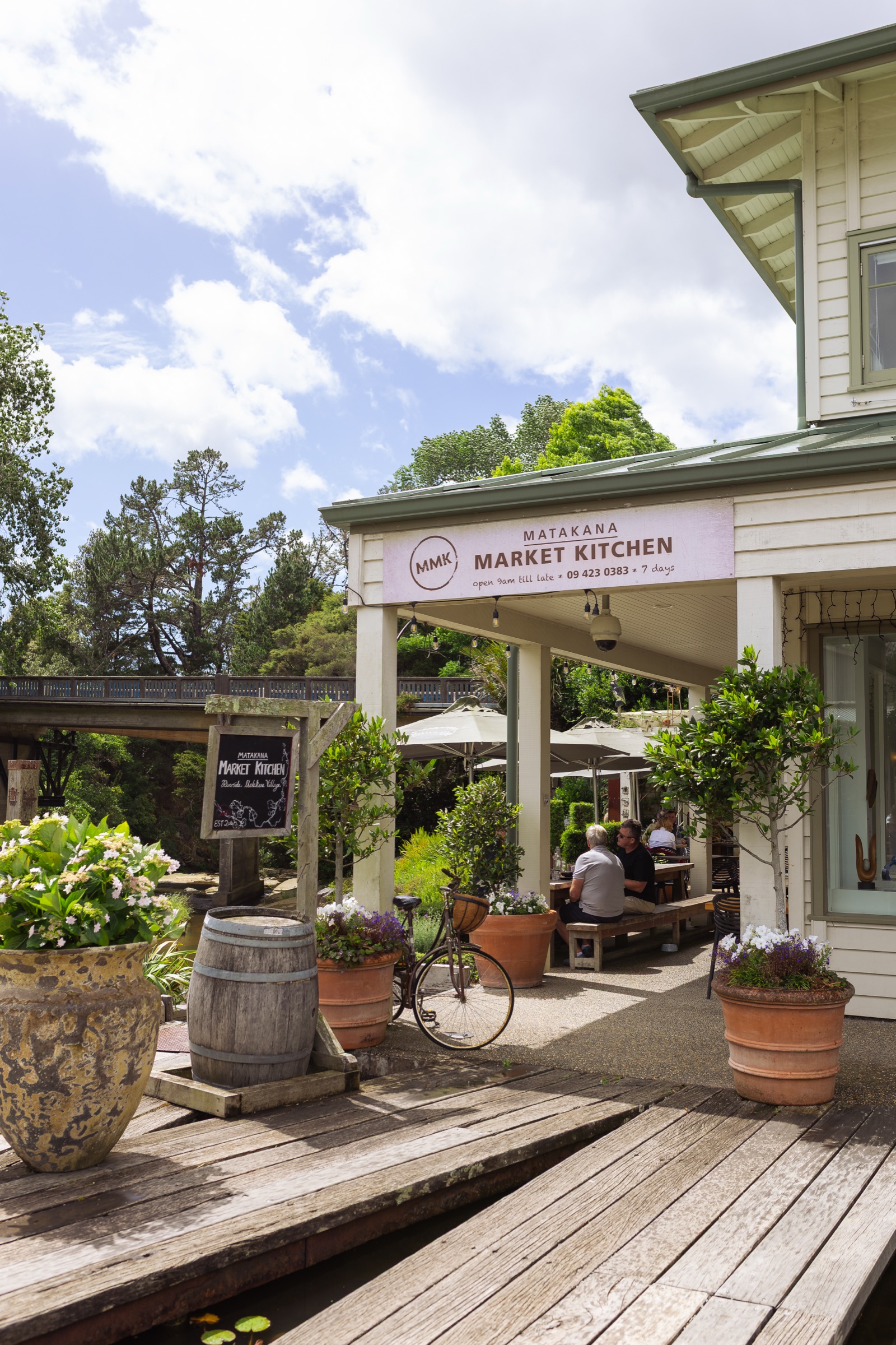 The entrance to Matakana Market Kitchen from the riverside deck: the sign over the verandah, a chalkboard, a wine barrel, terracotta pots of flowers, a bicycle, diners under umbrellas, and the village bridge behind.