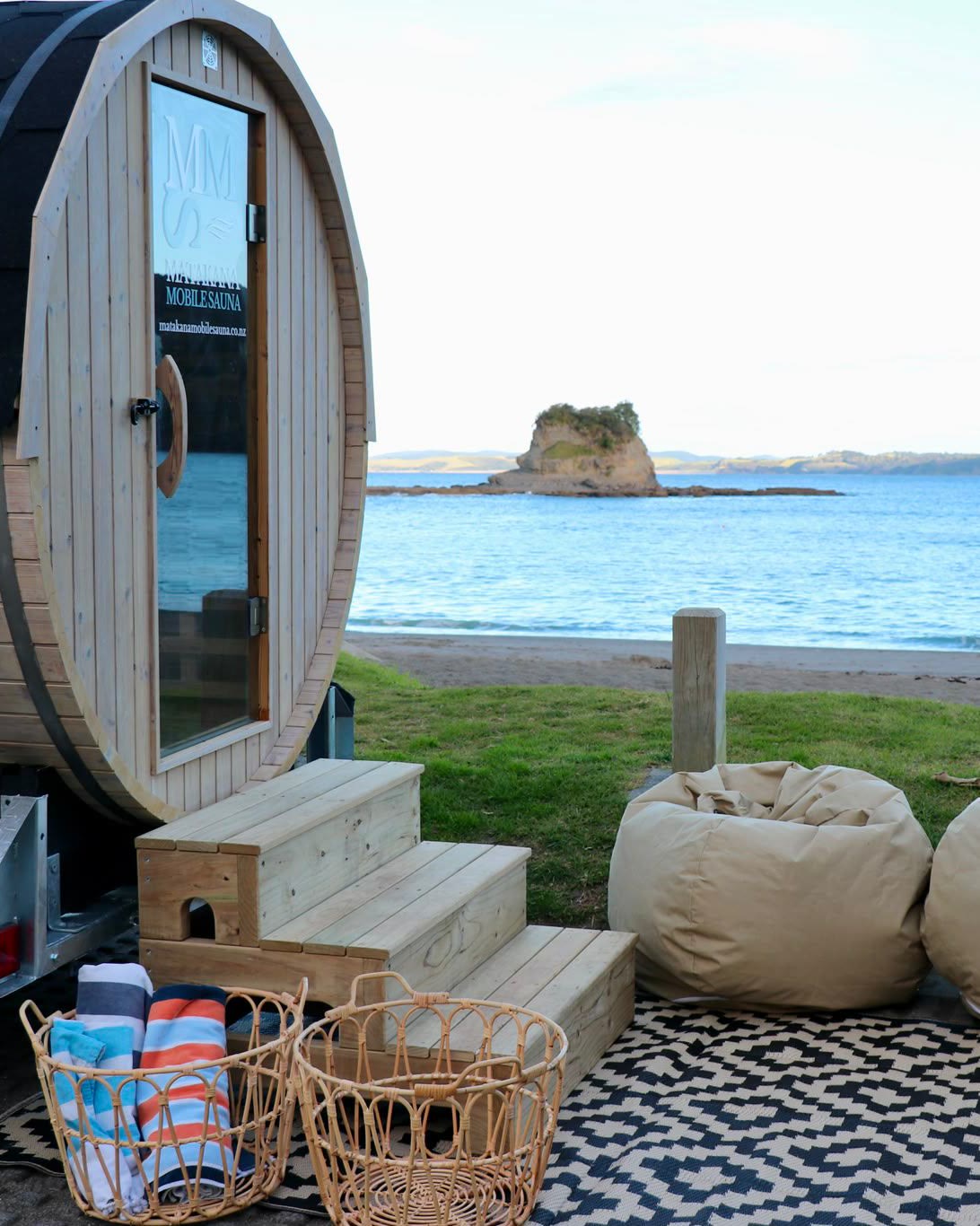 A timber barrel sauna on its trailer set up on the grass above a beach, steps to the glass door, two beanbags and baskets of striped towels on a patterned mat, and a rocky islet in the calm bay beyond.