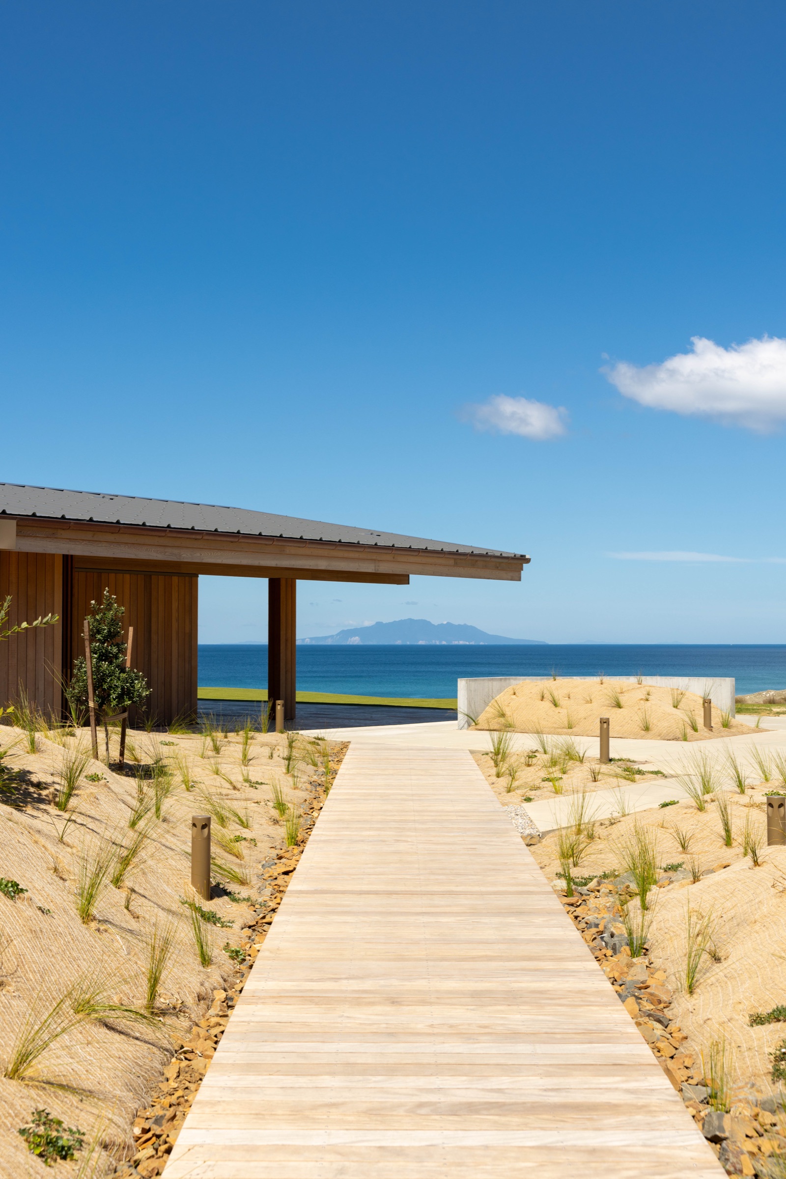 Ocean Restaurant at Te Arai Links: a timber boardwalk through planted dunes to the cedar pavilion, with the blue Pacific and the Hen and Chickens Islands beyond under a clear sky.