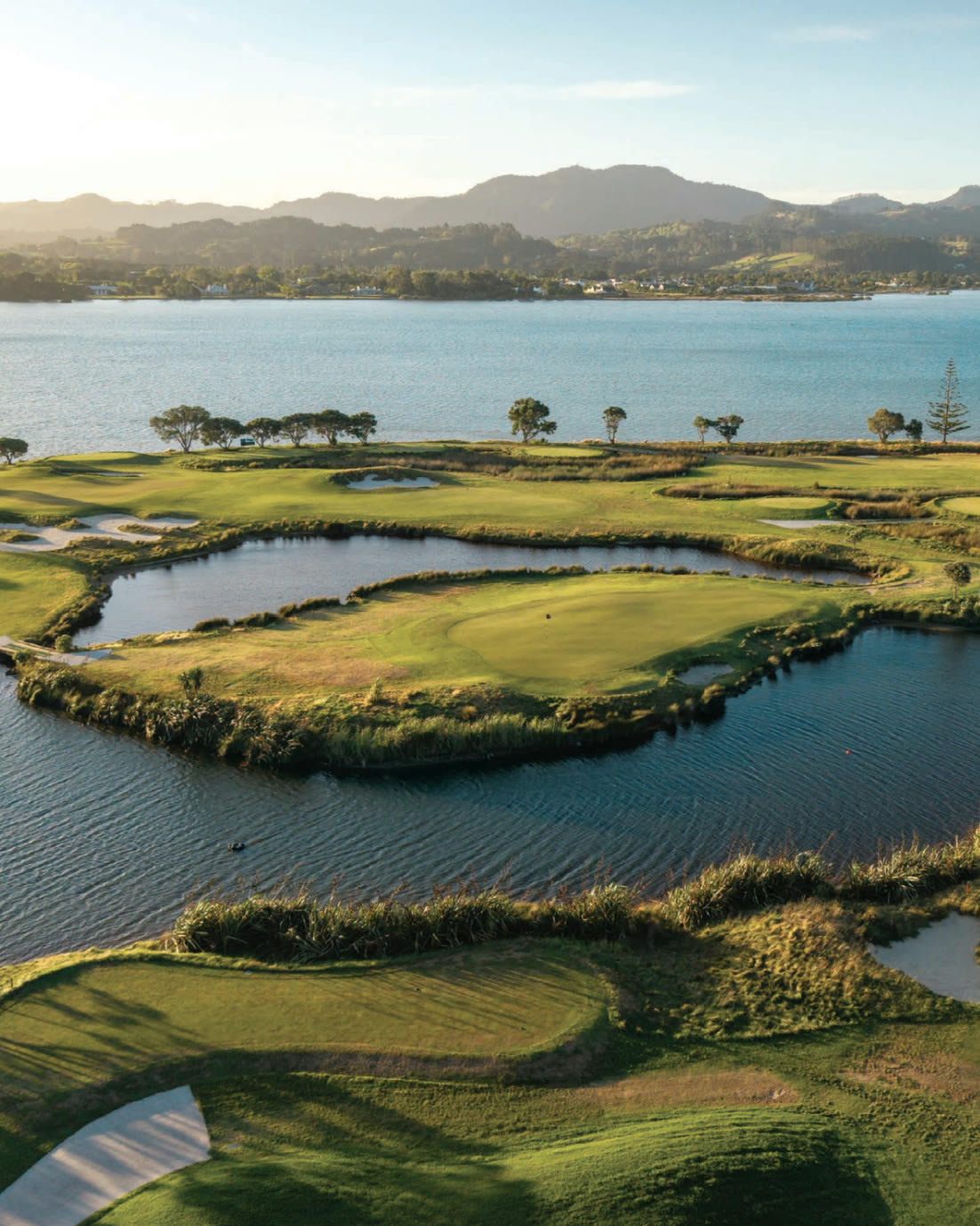 Omaha Beach Golf Club from the air at golden hour: the island green ringed by water and native grasses, bunkers and fairways beyond, the estuary and the hills behind.