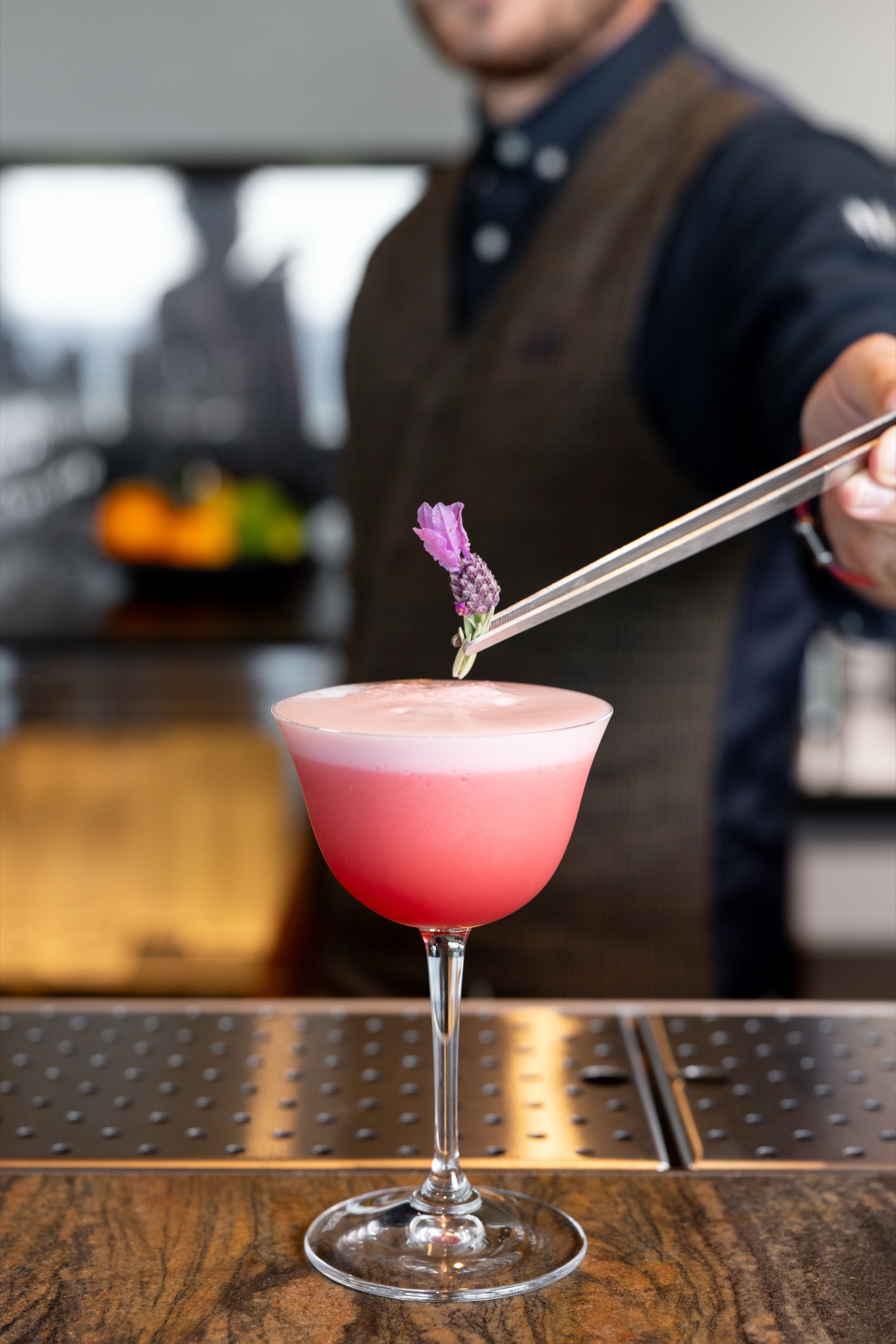 A bartender at Ric's at the Ocean, Te Arai Links, placing a sprig of lavender with tweezers onto a pink cocktail in a coupe on the bar, face out of frame.