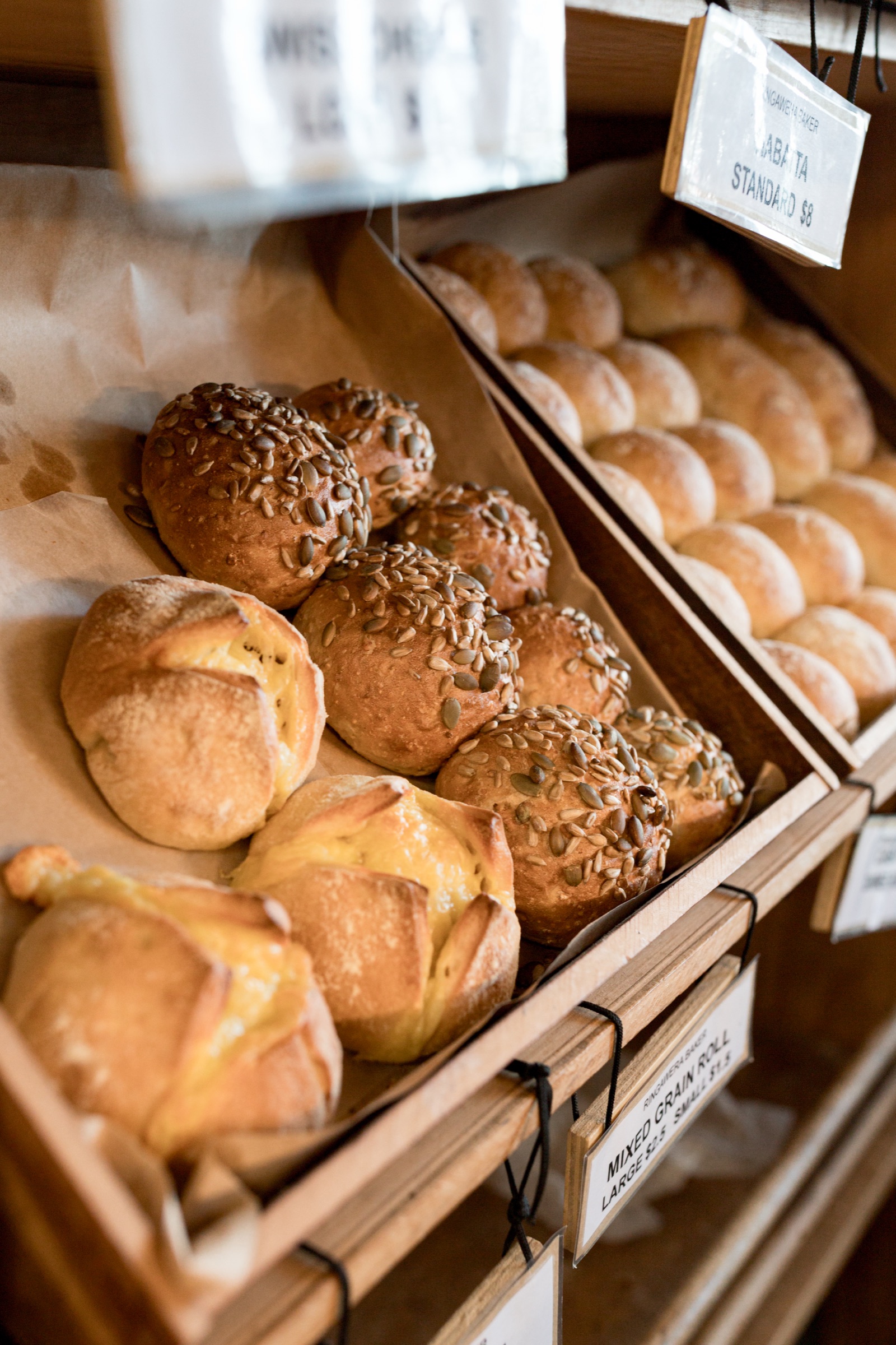 Bread at Rīngawera Baker, Matakana: seeded mixed grain rolls and cheese-topped buns in a wooden crate, ciabatta lined up in the crate behind, handwritten price tags on string.