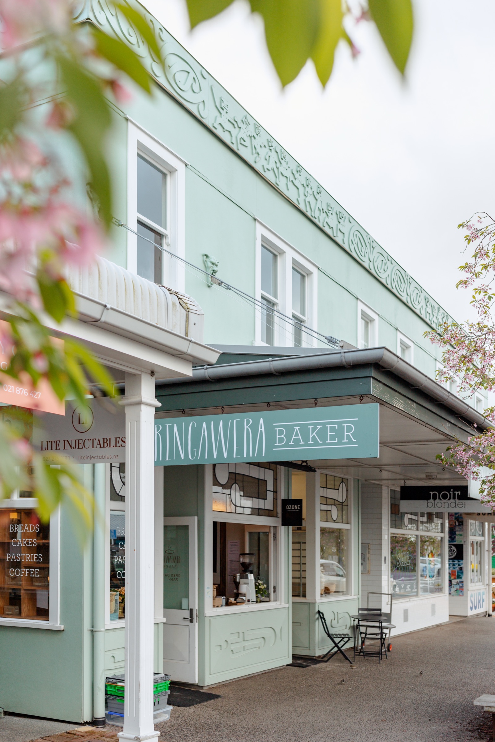 The Rīngawera Baker shopfront in Matakana Village: a sage green sign under the verandah of the ornate mint-green building, breads, cakes, pastries, and coffee lettered on the window, cherry blossom in the foreground.