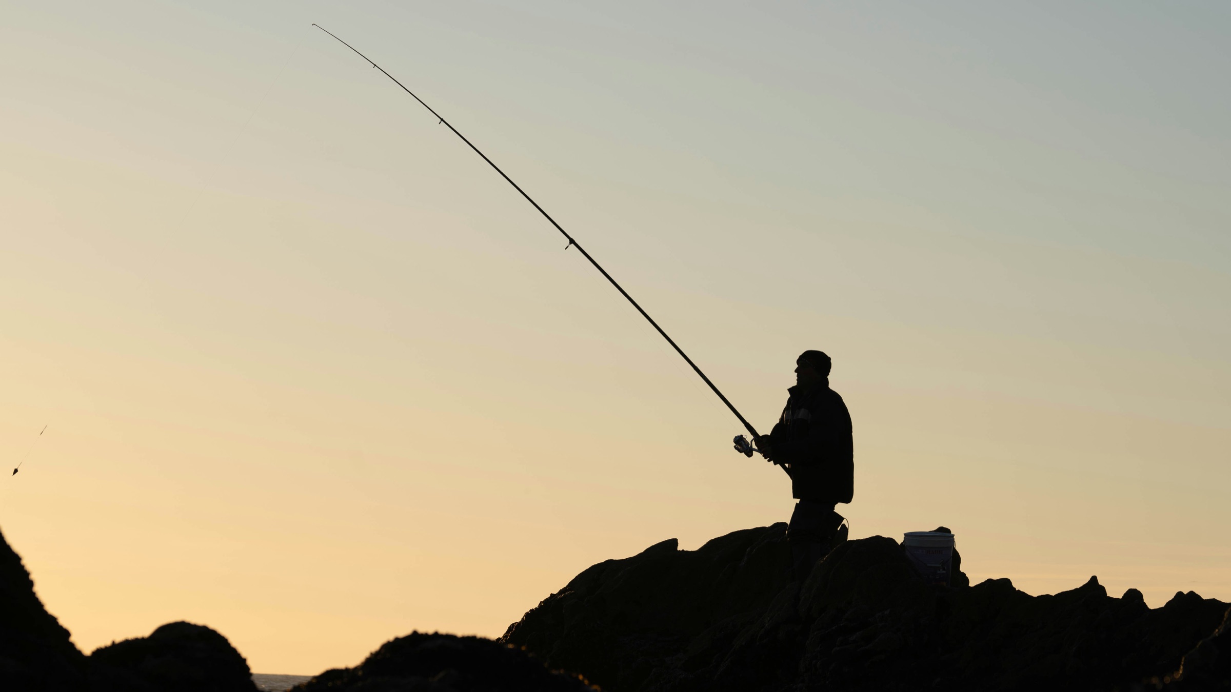 An angler in silhouette on the rocks at dusk, rod bent into the sea, the sky pale gold behind: fishing off the rocks.
