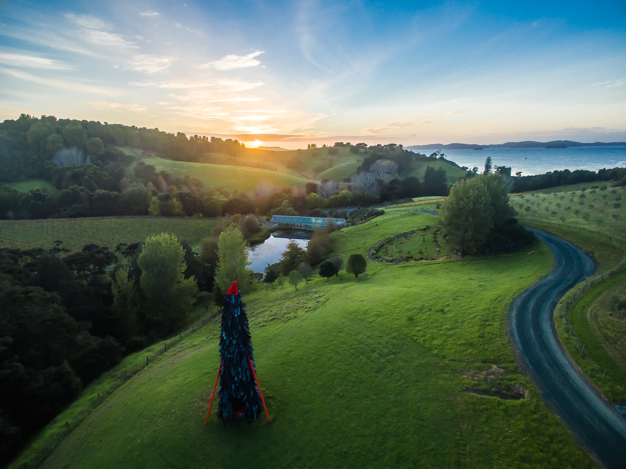 Brick Bay from the air at sunset: a tall feathered sculpture on the lawn, the glass house over the lake, vineyard rows, and Kawau Bay beyond, on a Terra and Tide Matakana Coast tour.