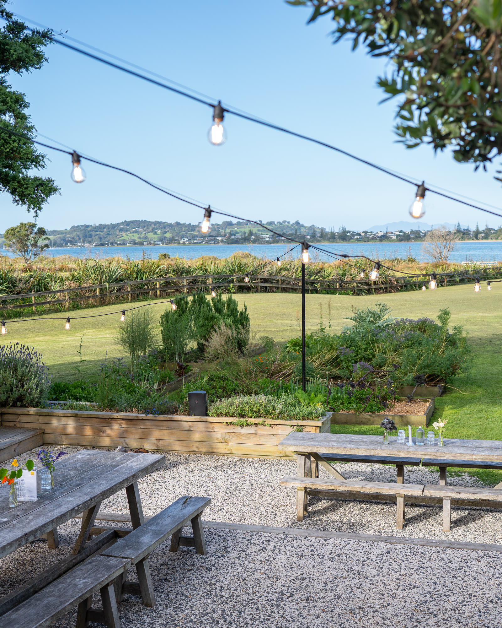 The garden at The Farmers Daughter, Omaha: timber picnic tables on gravel under festoon lights, raised beds of herbs and lavender, the lawn running to the estuary and the hills beyond.