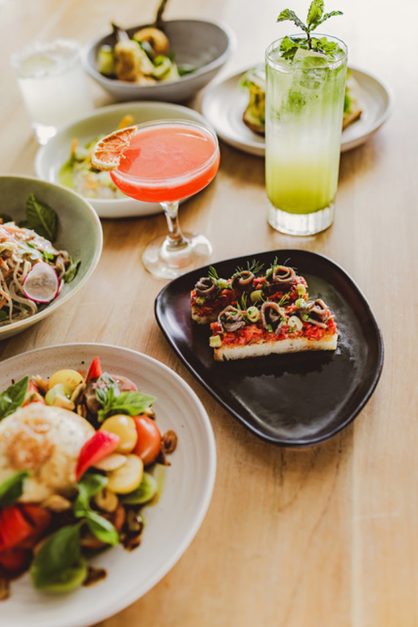 Summer plates at The Farmers Daughter, Omaha: anchovy and tomato toast on a black plate, a burrata and tomato salad, a noodle salad with radish, and two cocktails on a pale timber table.