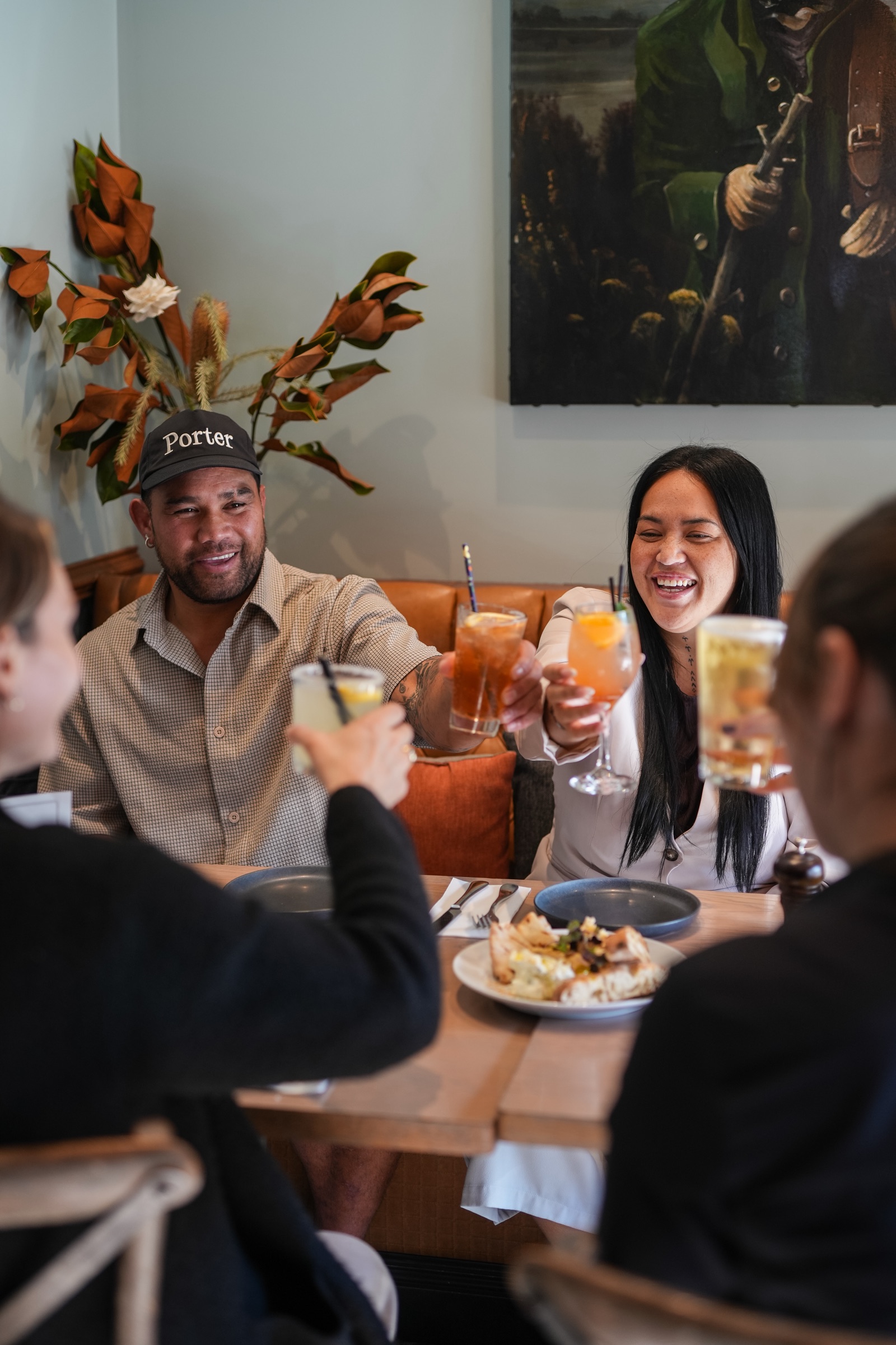 Four friends raising cocktails across a table in a leather booth at The Matakana Village Pub, a flatbread between them, dried magnolia in a vase and a painting on the wall behind.