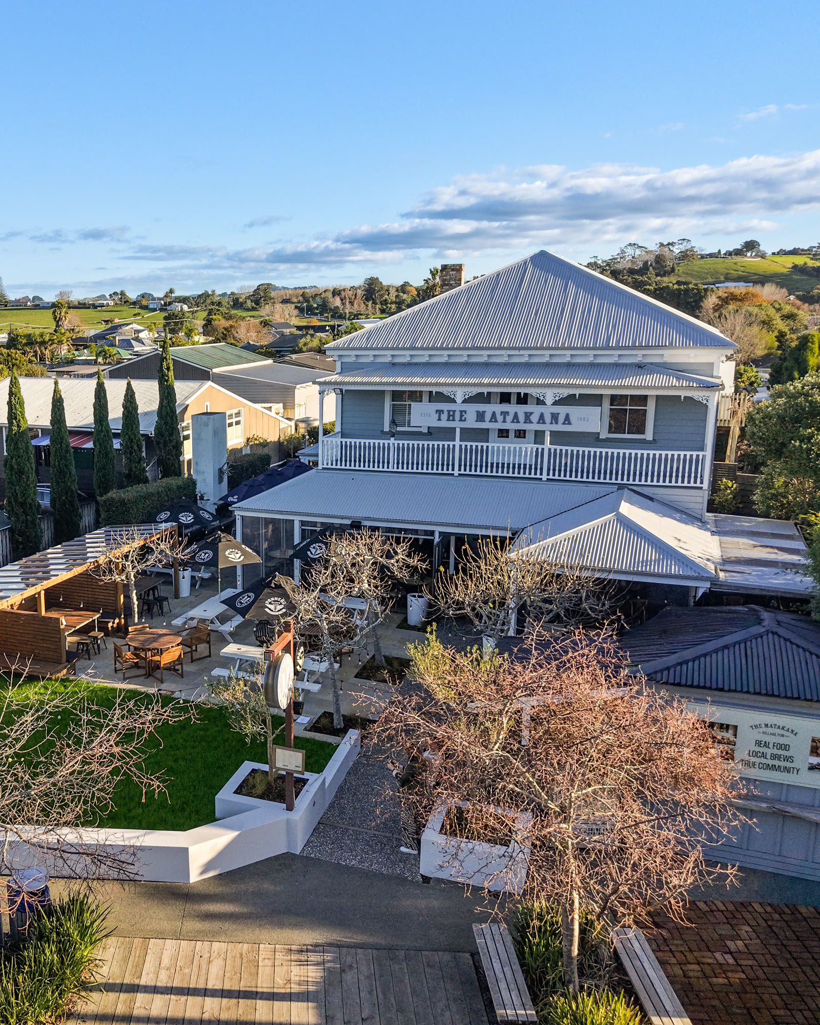 The Matakana Village Pub from the air: the white weatherboard hotel with its verandah and sign, the garden bar with pergola and tables on the lawn in front, cypress trees behind.