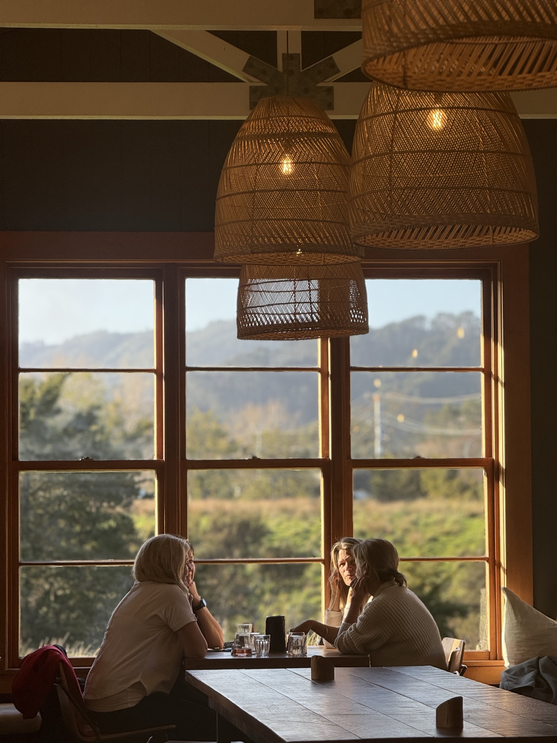 Inside the Smoko Room at Sawmill Brewery in evening light: three friends at a table by the tall timber-framed windows, rattan pendants glowing above, and the paddocks and hills beyond the glass.