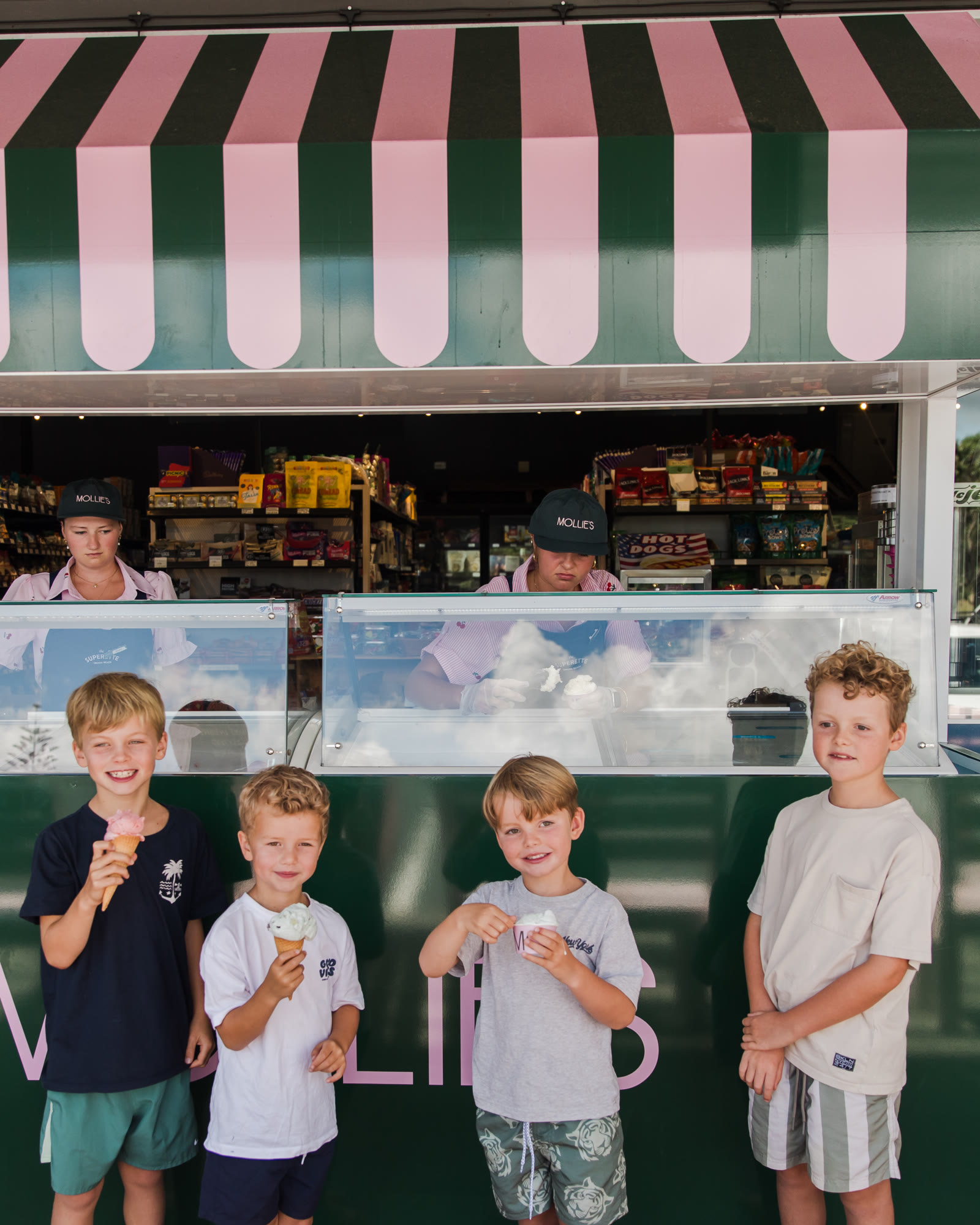 Four children with ice cream cones in front of the green counter at The Superette, Omaha Beach, staff scooping behind the glass under the striped awning.
