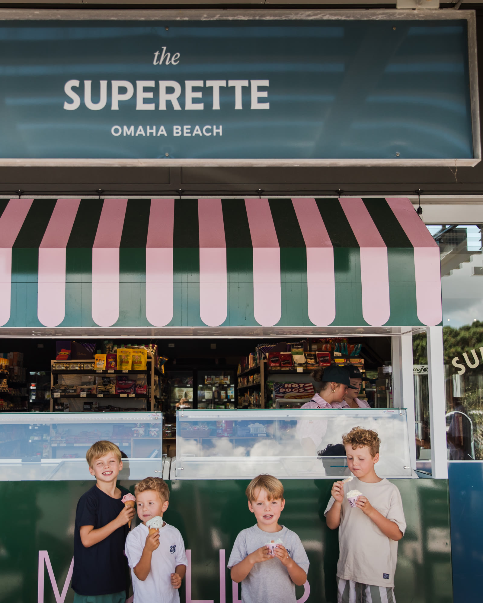 The Superette at Omaha Beach: the shop sign above the pink and green striped awning of the ice cream counter, four children with cones in front of the green counter, and staff scooping behind the glass.