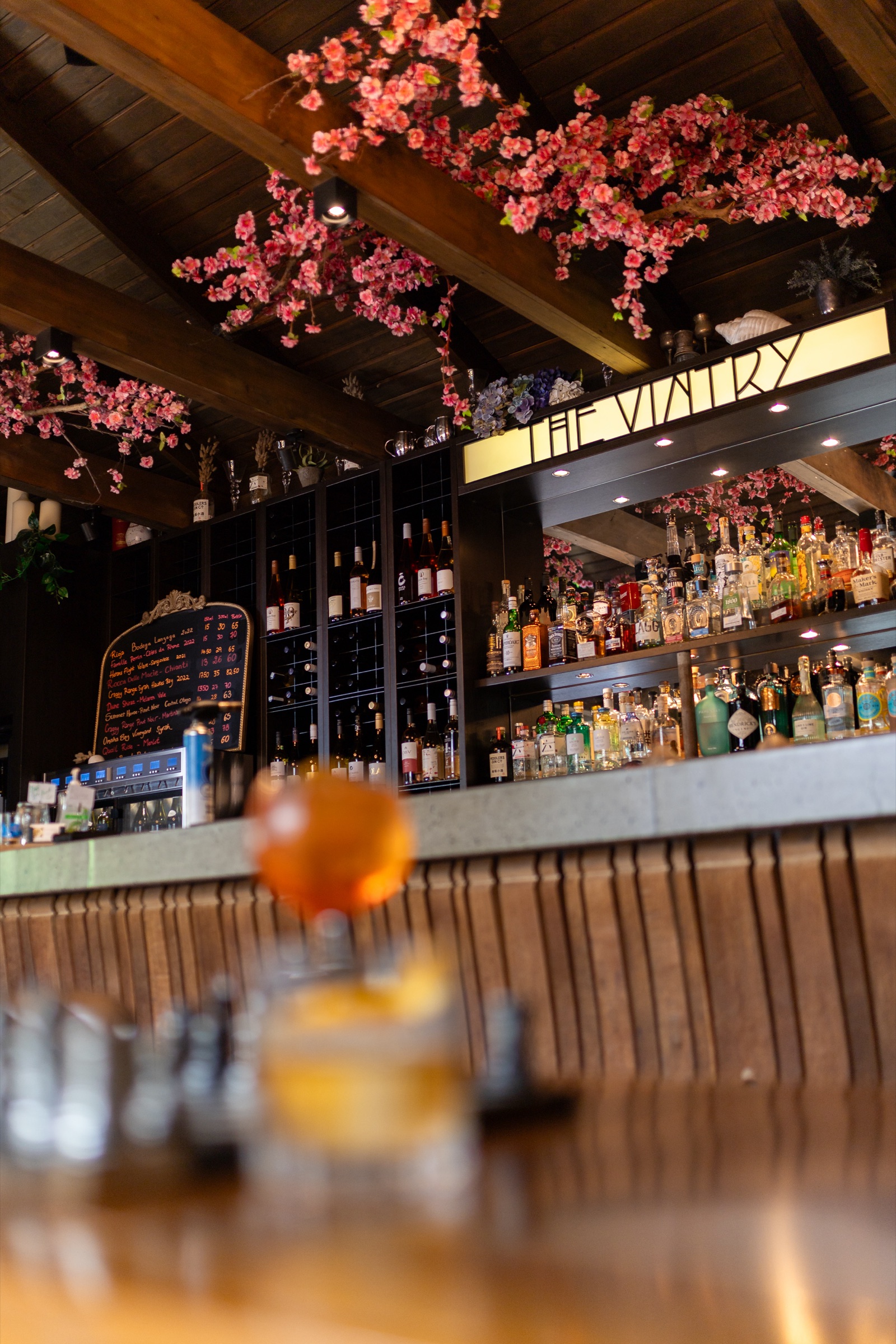 The bar at The Vintry, Matakana: the lit sign above shelves of local wine and spirits, pink blossom along the timber beams, a chalkboard wine list, and a glass of rosé on the counter in front.