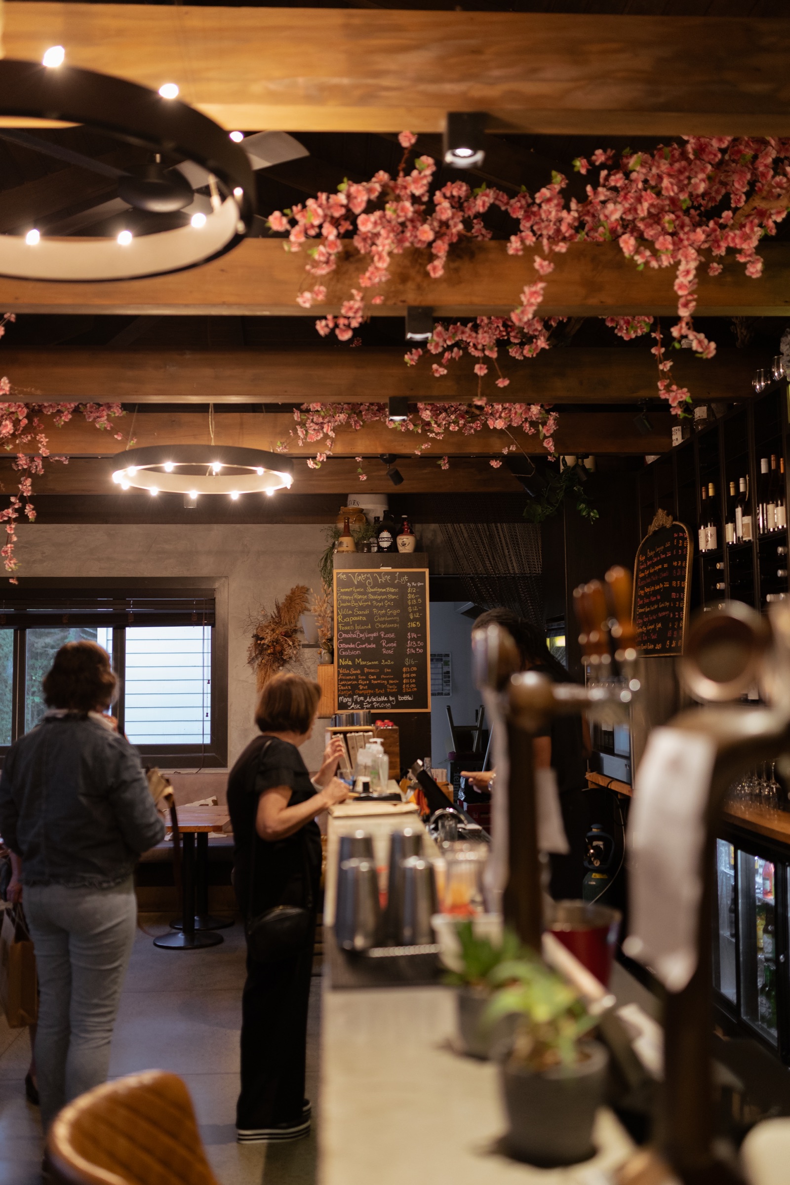 Inside The Vintry, Matakana: guests at the bar beneath ring lights and blossom-hung beams, the chalkboard wine list of local pours behind, and the taps in the foreground.