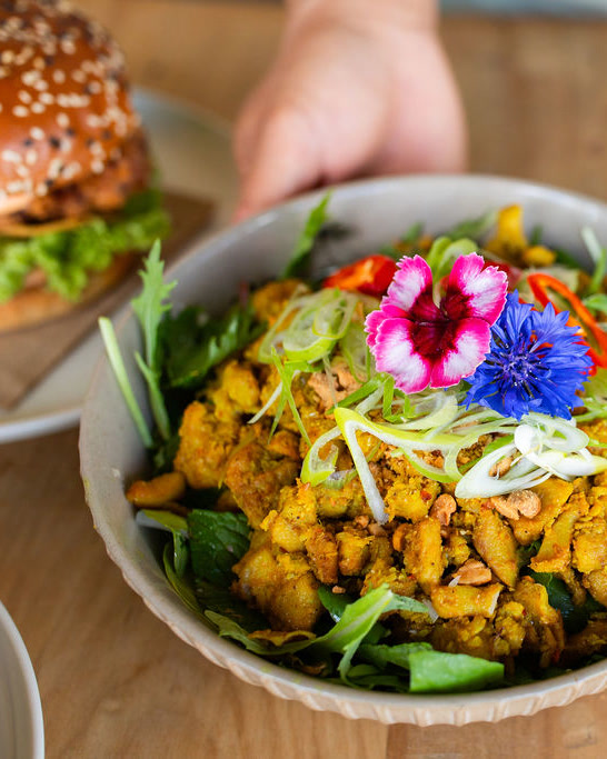 A bowl of crispy turmeric chicken salad topped with spring onion and edible flowers at Viet Q, Matakana, held over the timber table, with a sesame-bun burger on a plate beside it.
